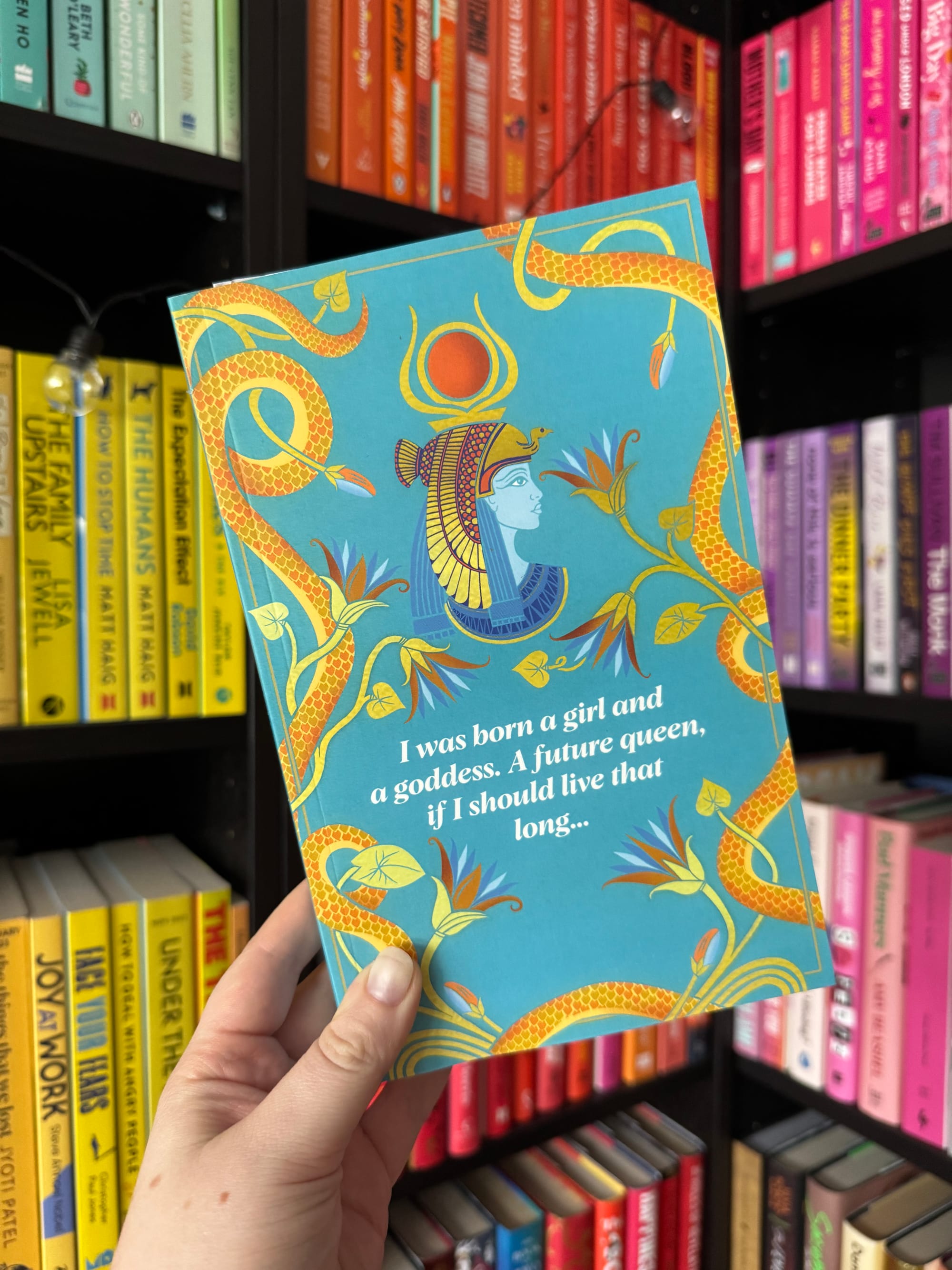 Hand holds a blue book with golden snake designs and an Egyptian motif. Text reads: "I was born a girl and a goddess..." Colorful bookshelves in back.