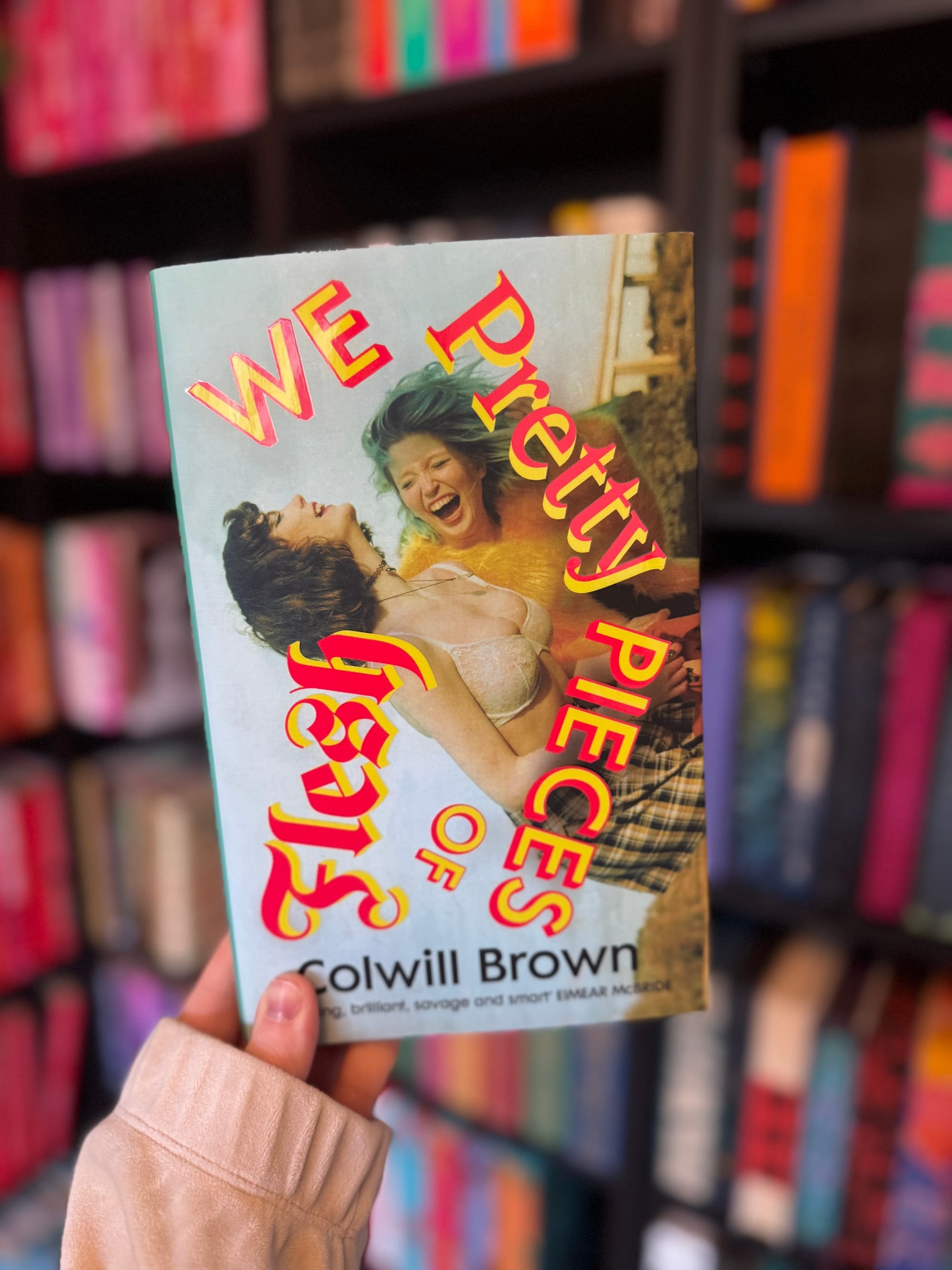 Hand holding "Pretty Pieces of Flesh" book with colorful cover showing two laughing women. Bookshelves with various colored books in background.
