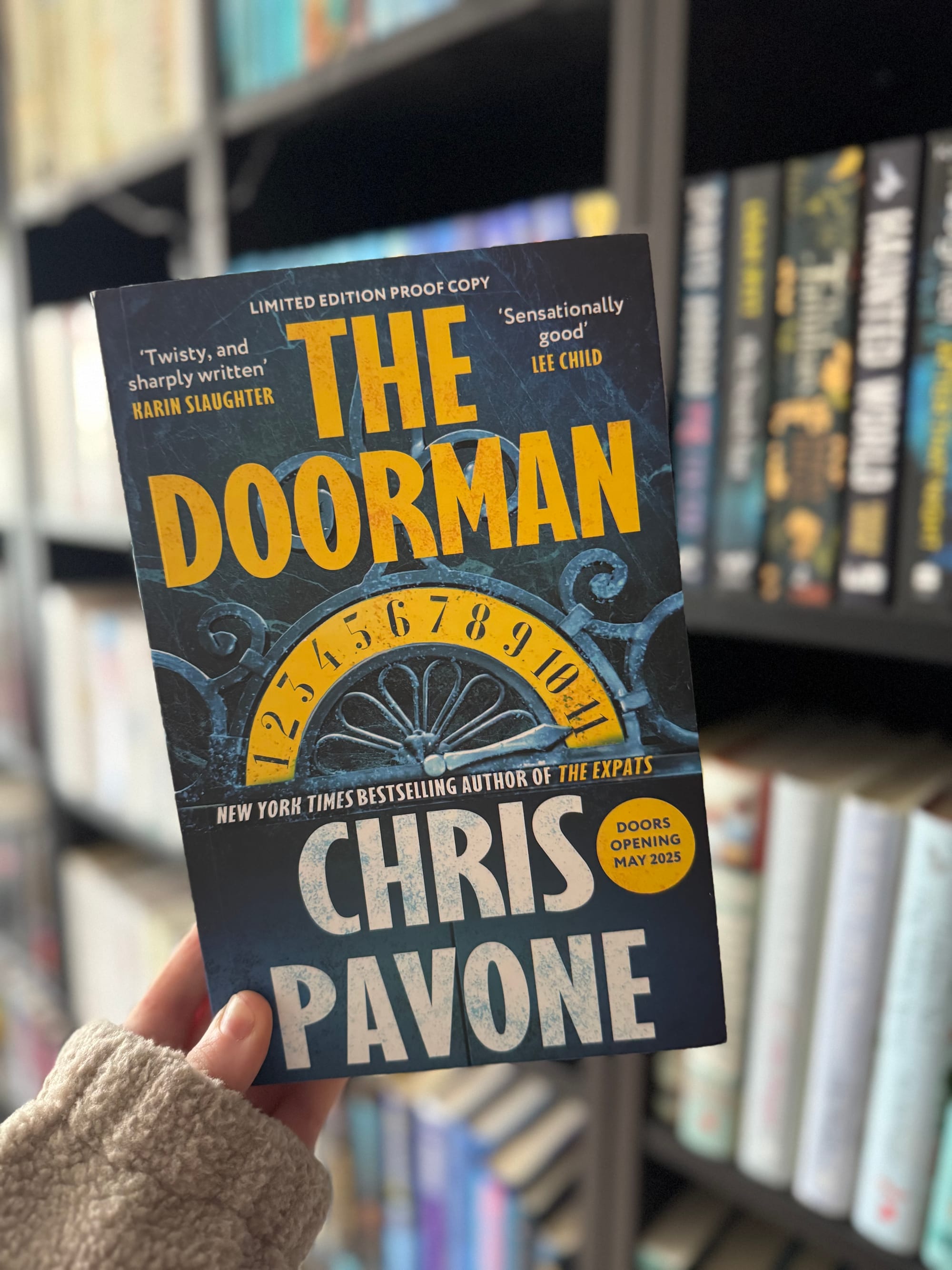 A hand holds "The Doorman" by Chris Pavone against a blurred bookshelf. The cover features a clock design and quotes from authors.