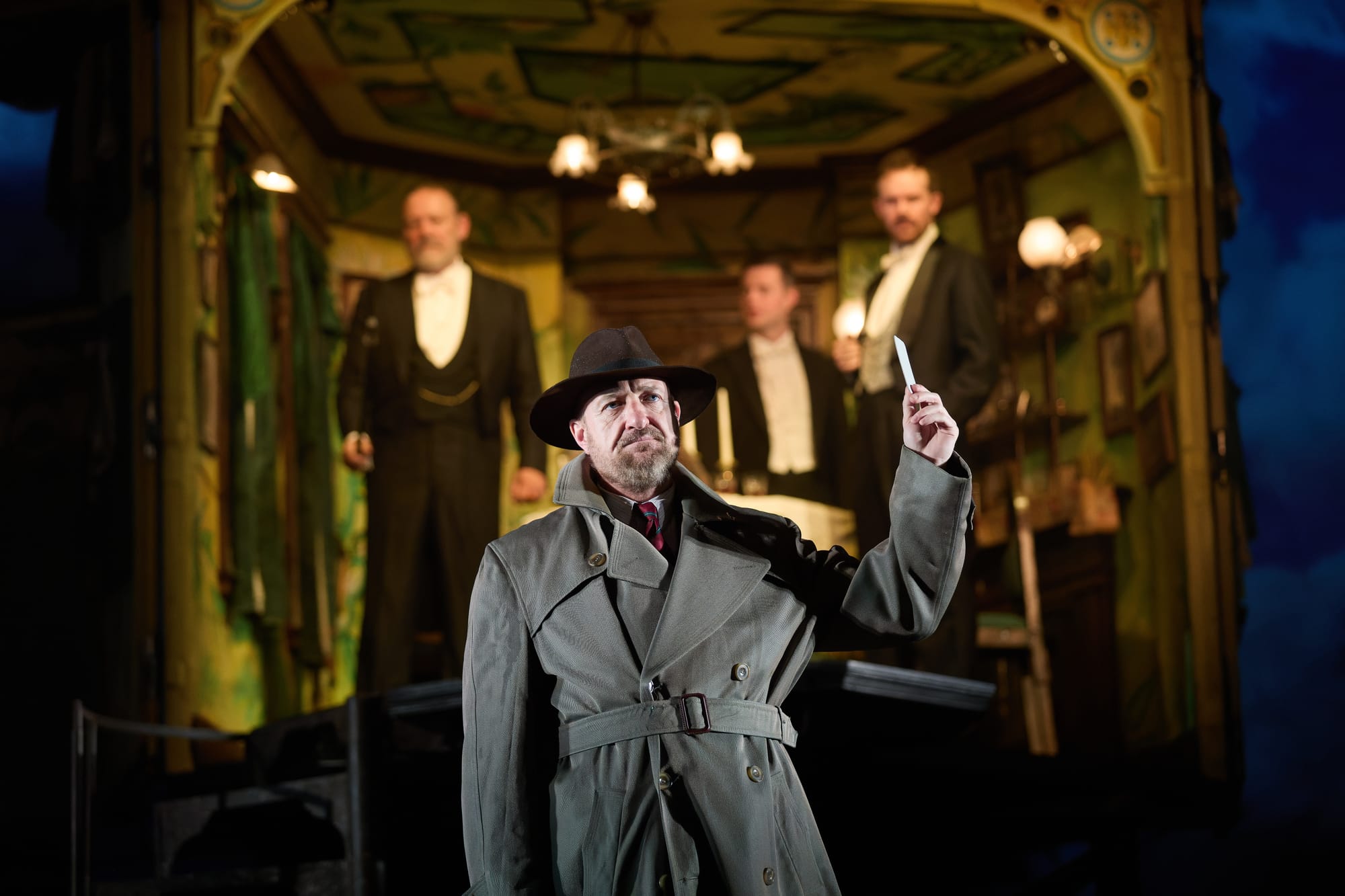Man in trench coat and hat holds paper, standing in front of three men in suits inside a decorated room. Dim lighting and vintage decor.