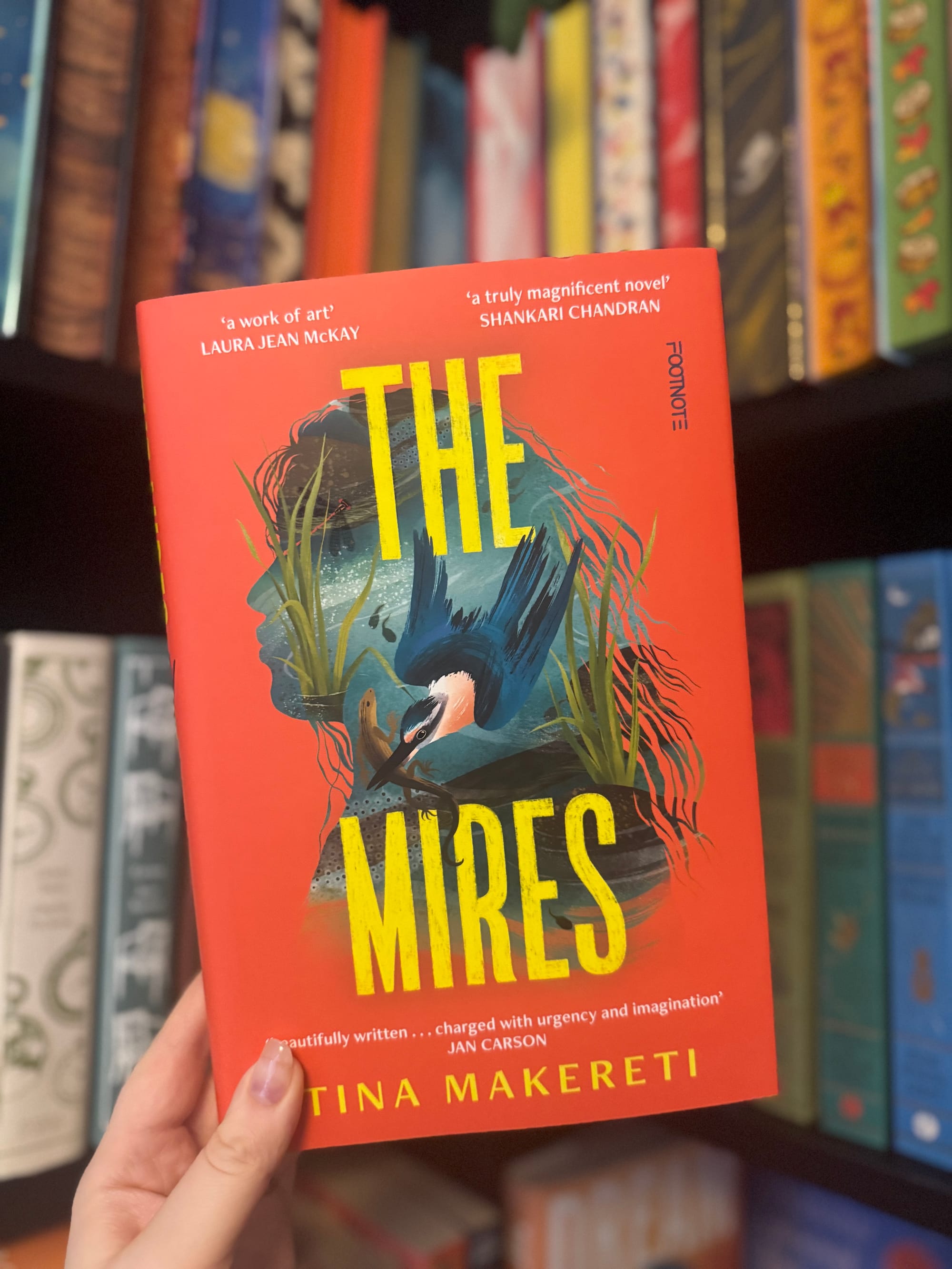 Person holding a red book titled "The Mires" by Tina Makereti. The cover features a bird and plants, with shelves of colorful books in the background.