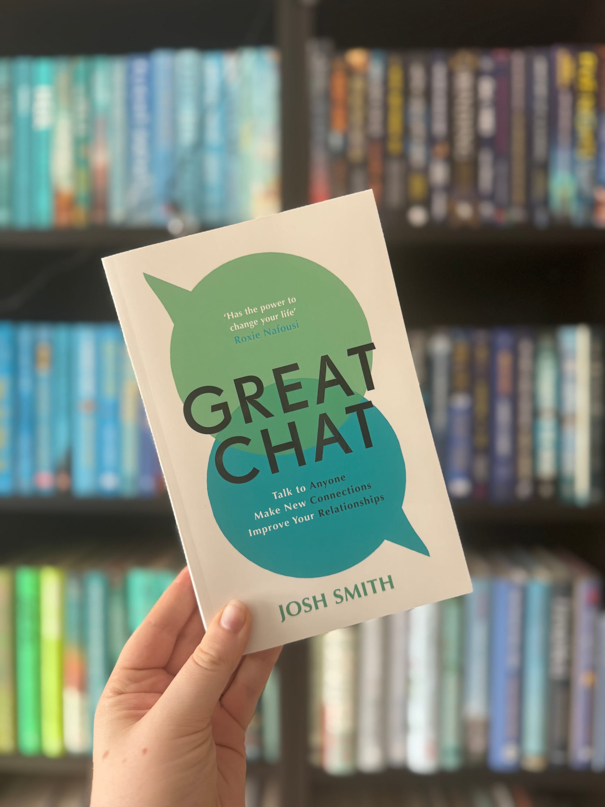 Hand holding the book "Great Chat" by Josh Smith, with a green speech bubble design. Bookshelves with colorful spines in the background.