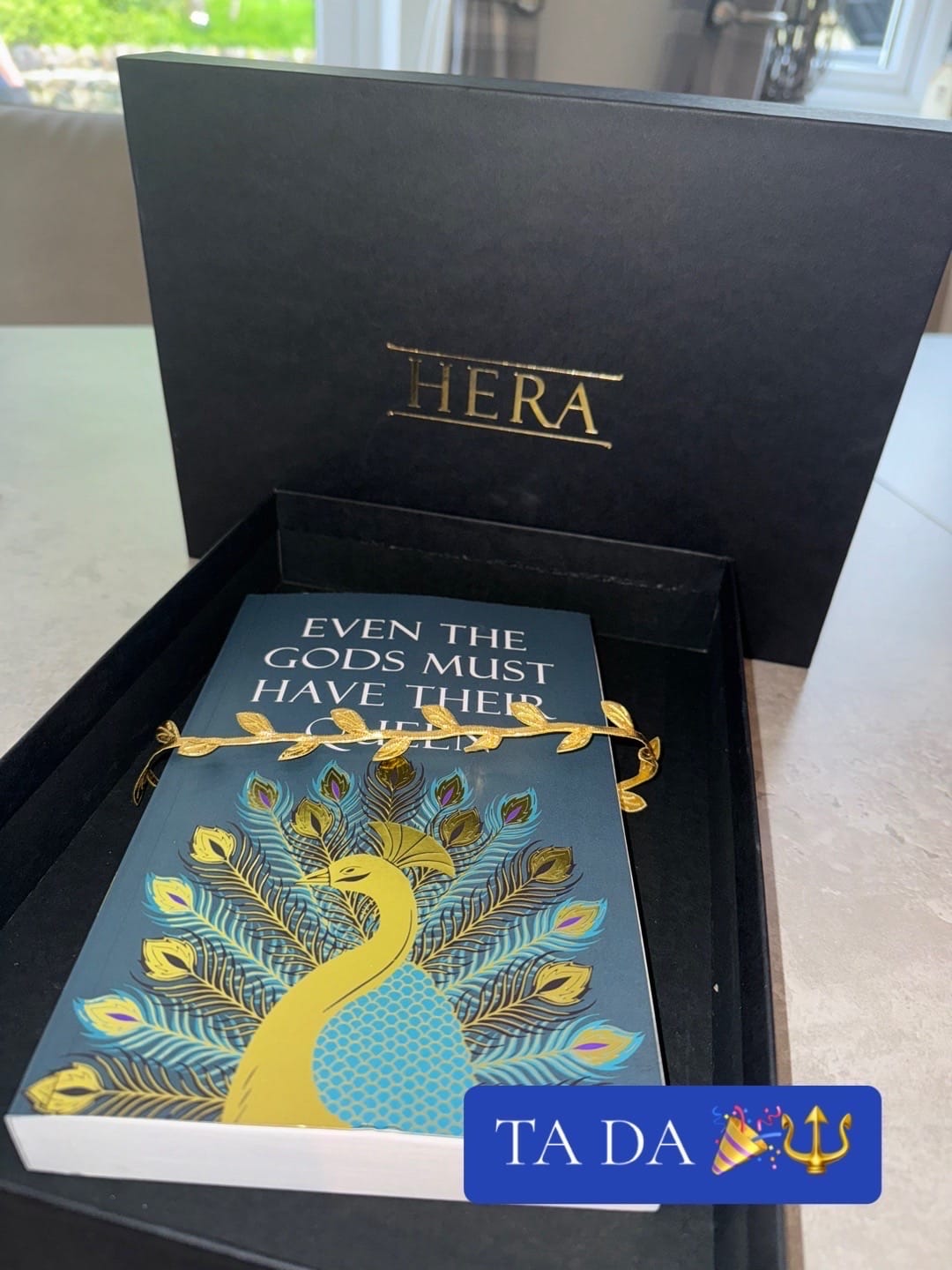 Box with "HERA" contains a book titled "EVEN THE GODS MUST HAVE THEIR..." adorned with a peacock design and gold laurel. Text reads "TA DA ππ±".
