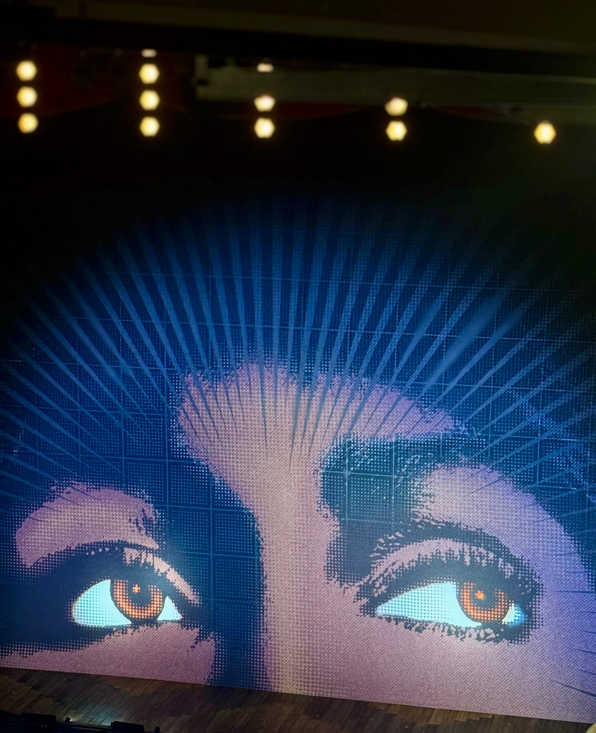Close-up artistic image of eyes with brown irises on a blue patterned background under stage lights. Mood is dramatic and intriguing.