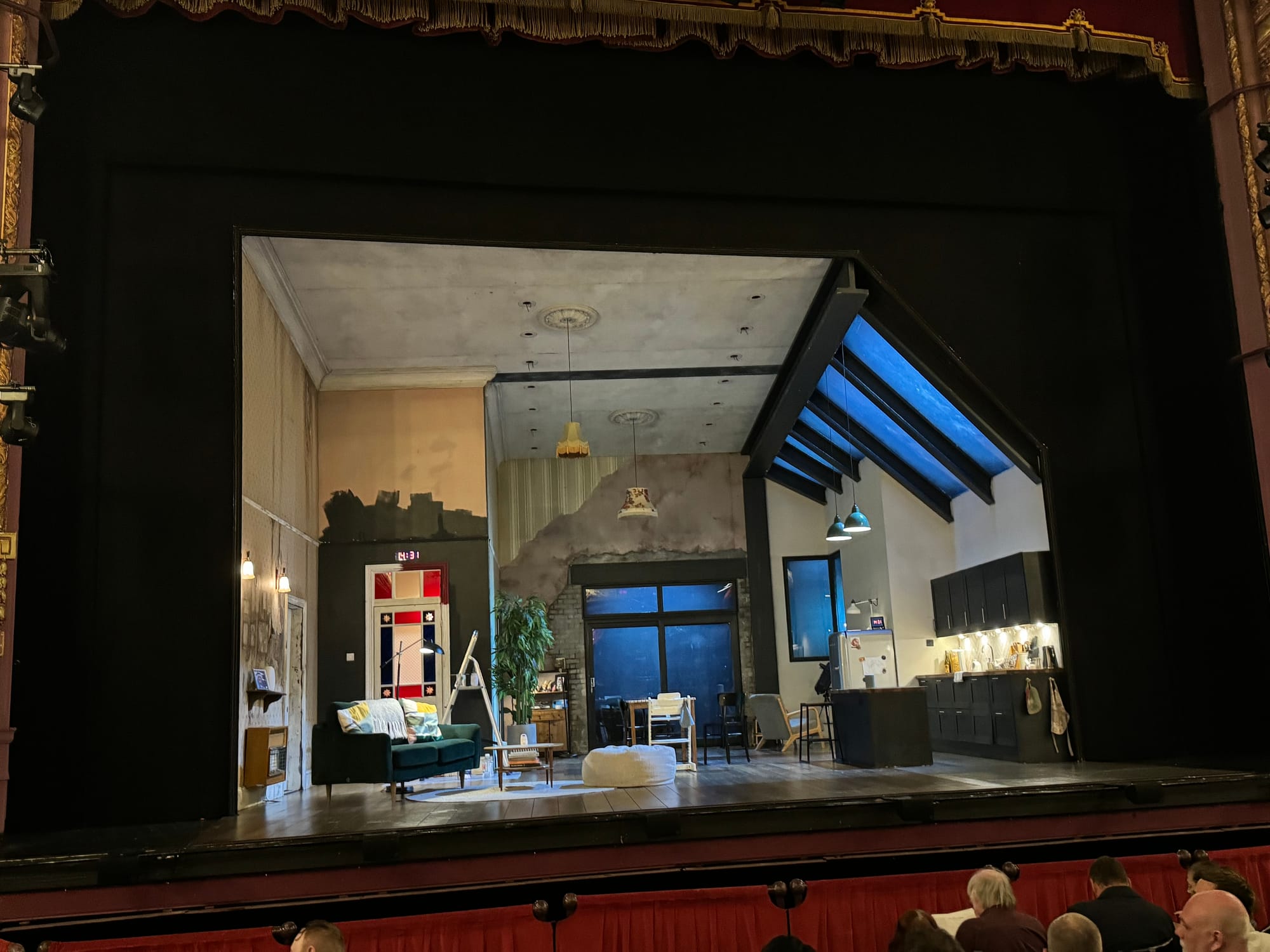 Theater stage set as a modern living room with a black sofa, dining area, and kitchen. Audience visible in front, creating an anticipatory mood.