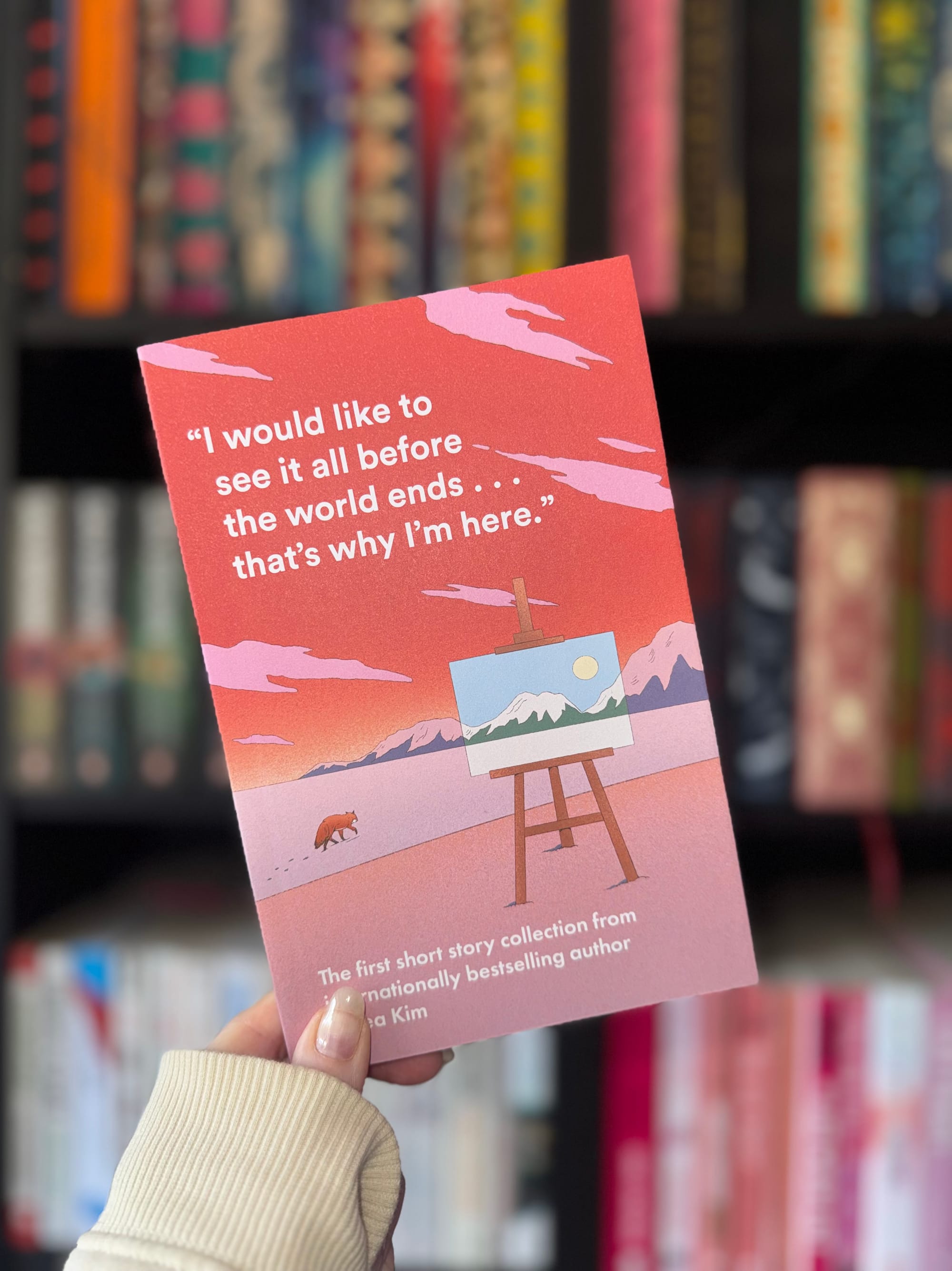 Hand holds a red book with a mountain and fox illustration. Quote says, "I would like to see it all before the world ends... that's why I'm here." Bookshelf blurred in background.