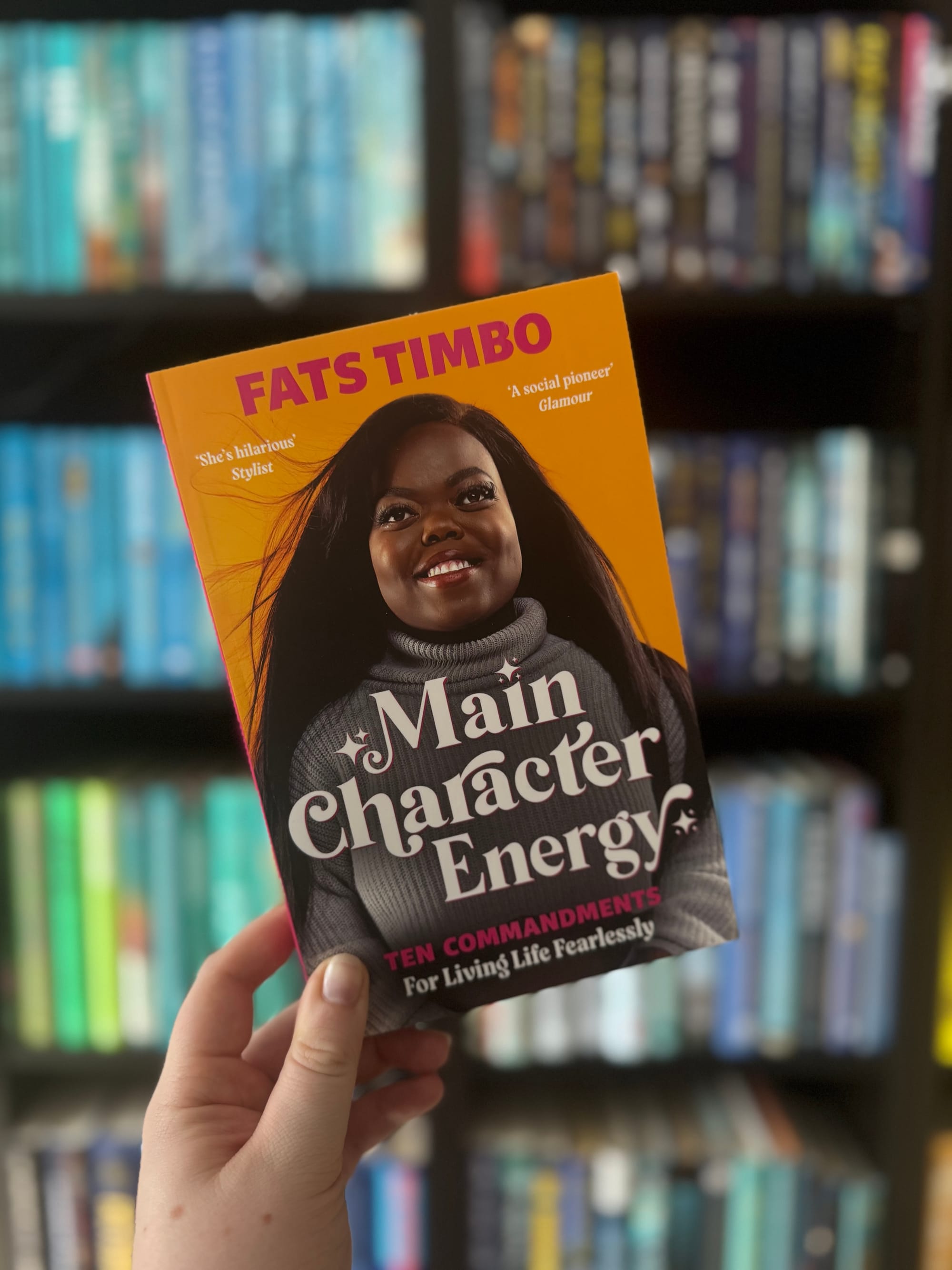 A hand holds a book titled "Main Character Energy" by Fats Timbo against a blurred bookshelf background. The cover is bright yellow-orange.