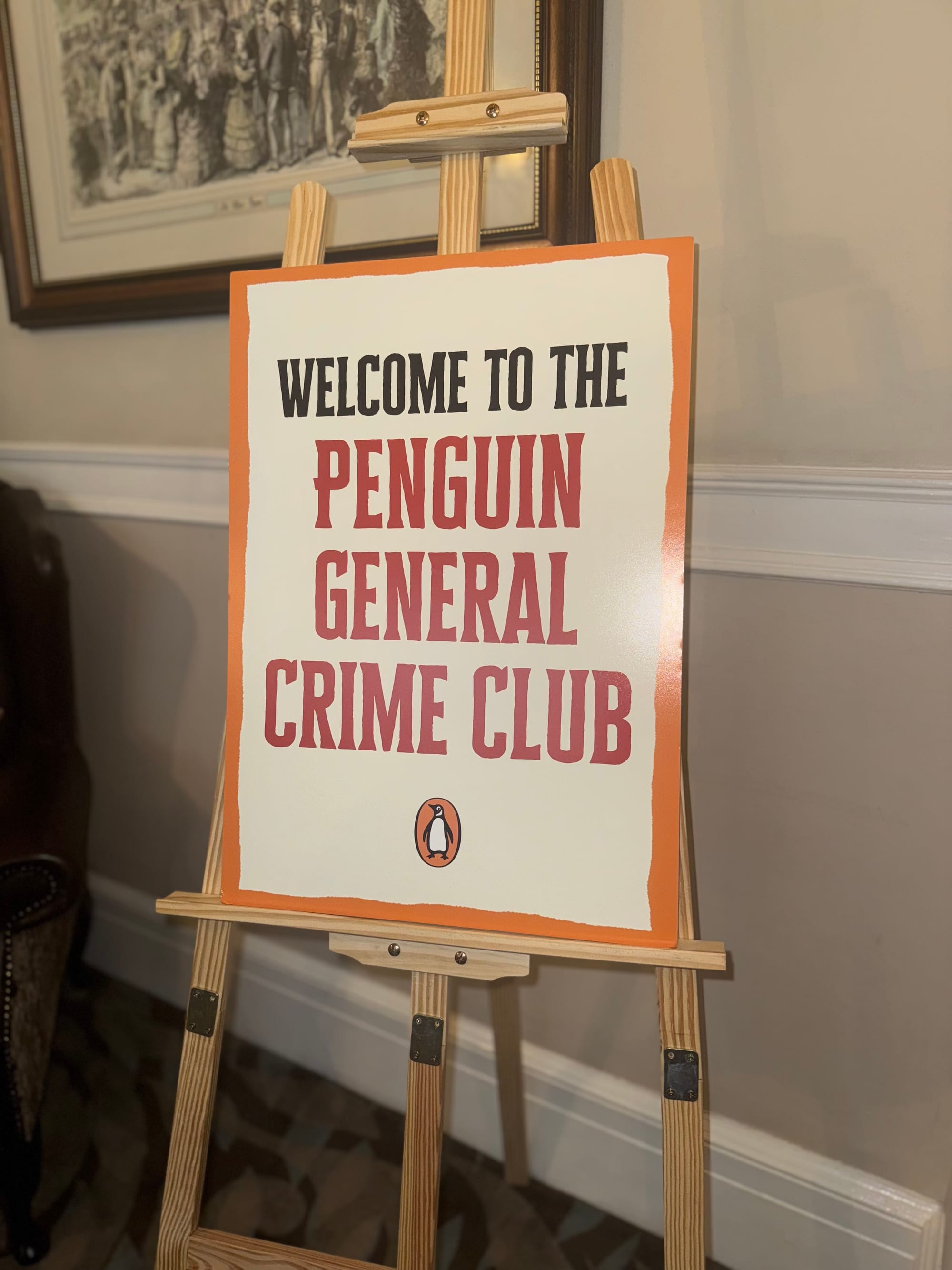 Easel display board reading “Welcome to the Penguin General Crime Club” with the Penguin logo, placed in a traditional room with framed artwork and soft lighting.