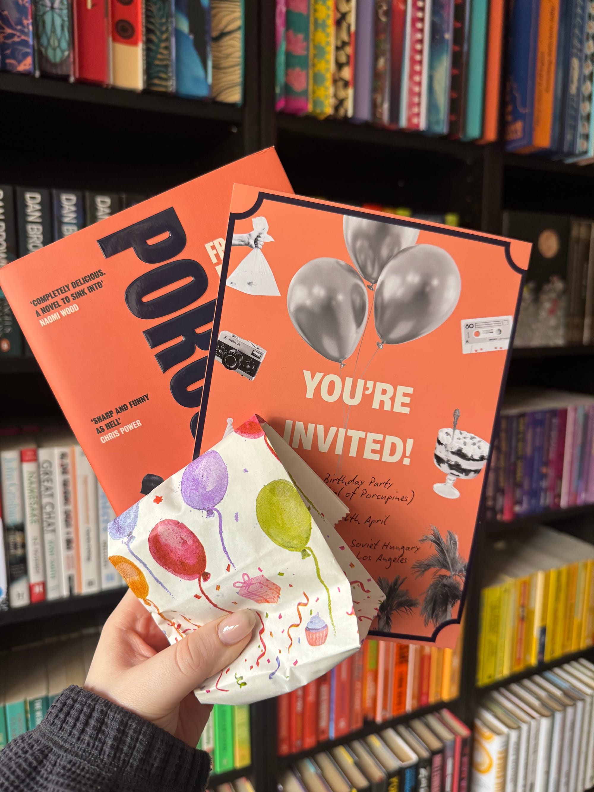 A hand holds orange books and a gift bag with balloons, set against colorful bookshelves. One book cover reads "You're Invited!"