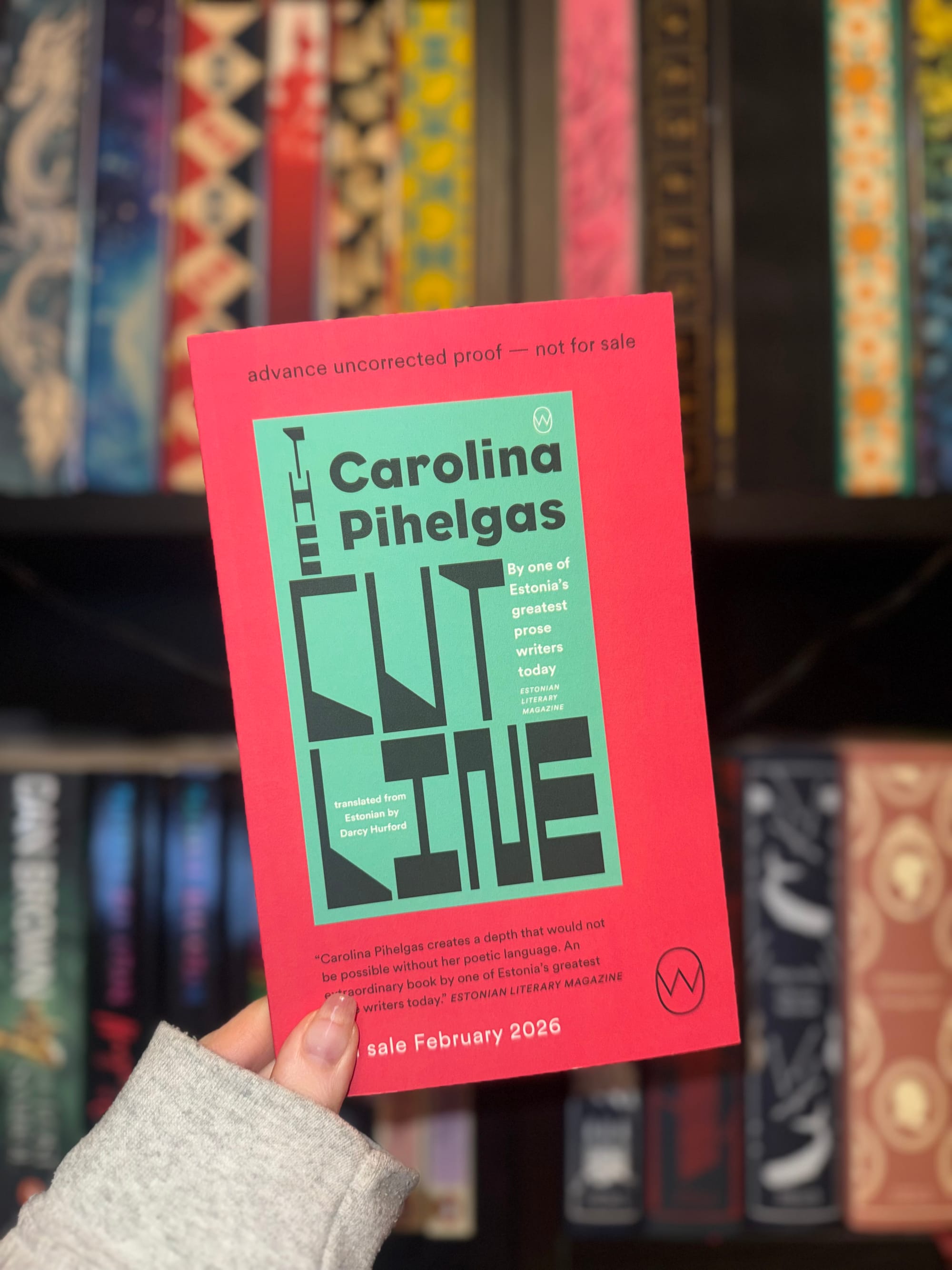 Hand holding a red and green book titled "The Cut Line" by Carolina Pihelgas, in front of a colorful patterned bookshelf.