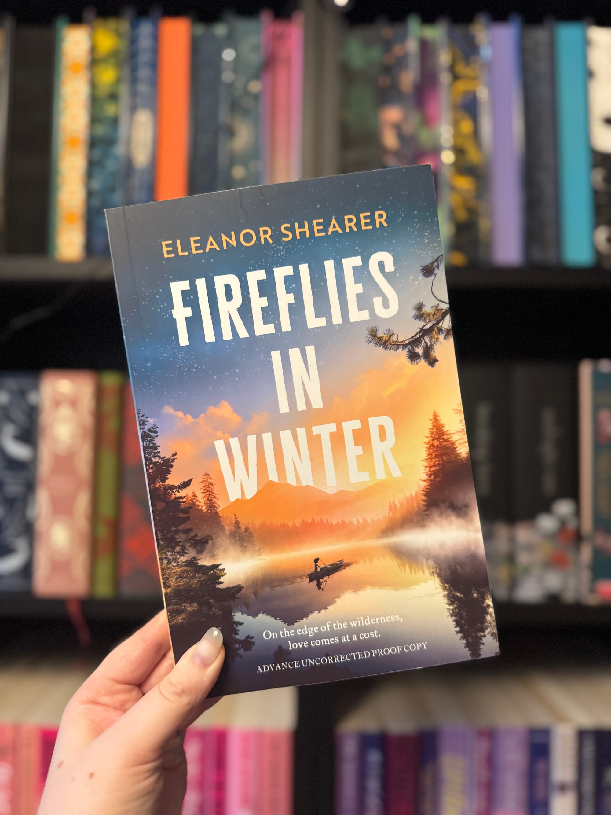 Hand holding "Fireflies in Winter" by Eleanor Shearer against a bookshelf. The cover shows a canoe on a misty lake at sunset.