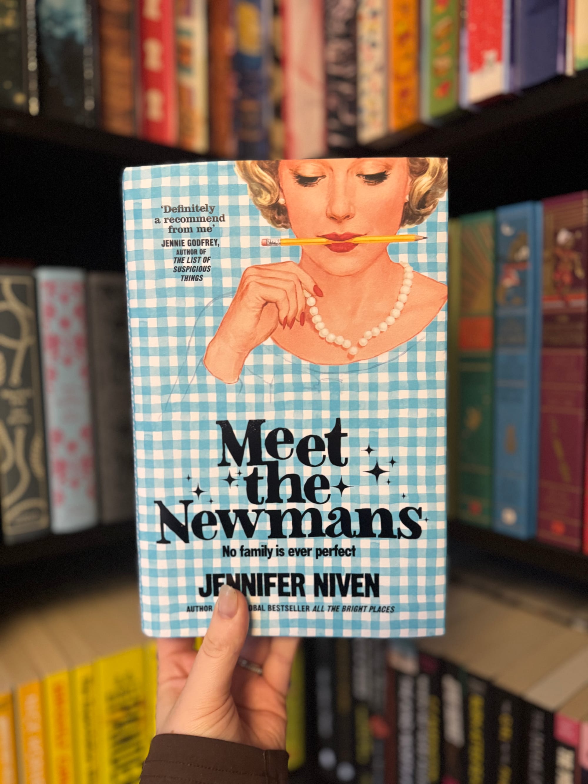 Hand holding "Meet the Newmans" book with blue checkered cover, showing a woman balancing a pencil. Shelves of colorful books in background.