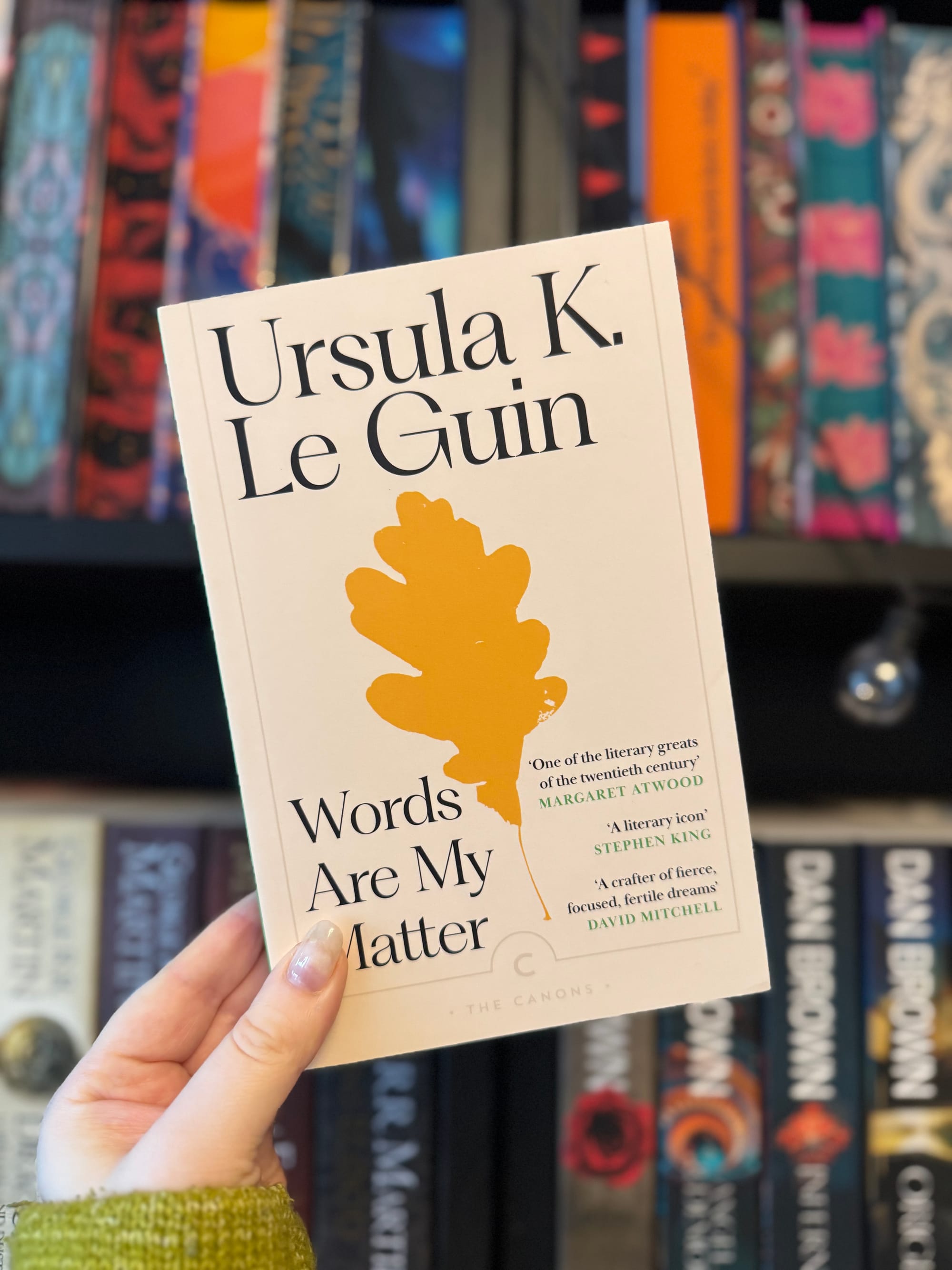 Hand holding "Words Are My Matter" book by Ursula K. Le Guin against colorful bookshelf. Cover features orange leaf design.