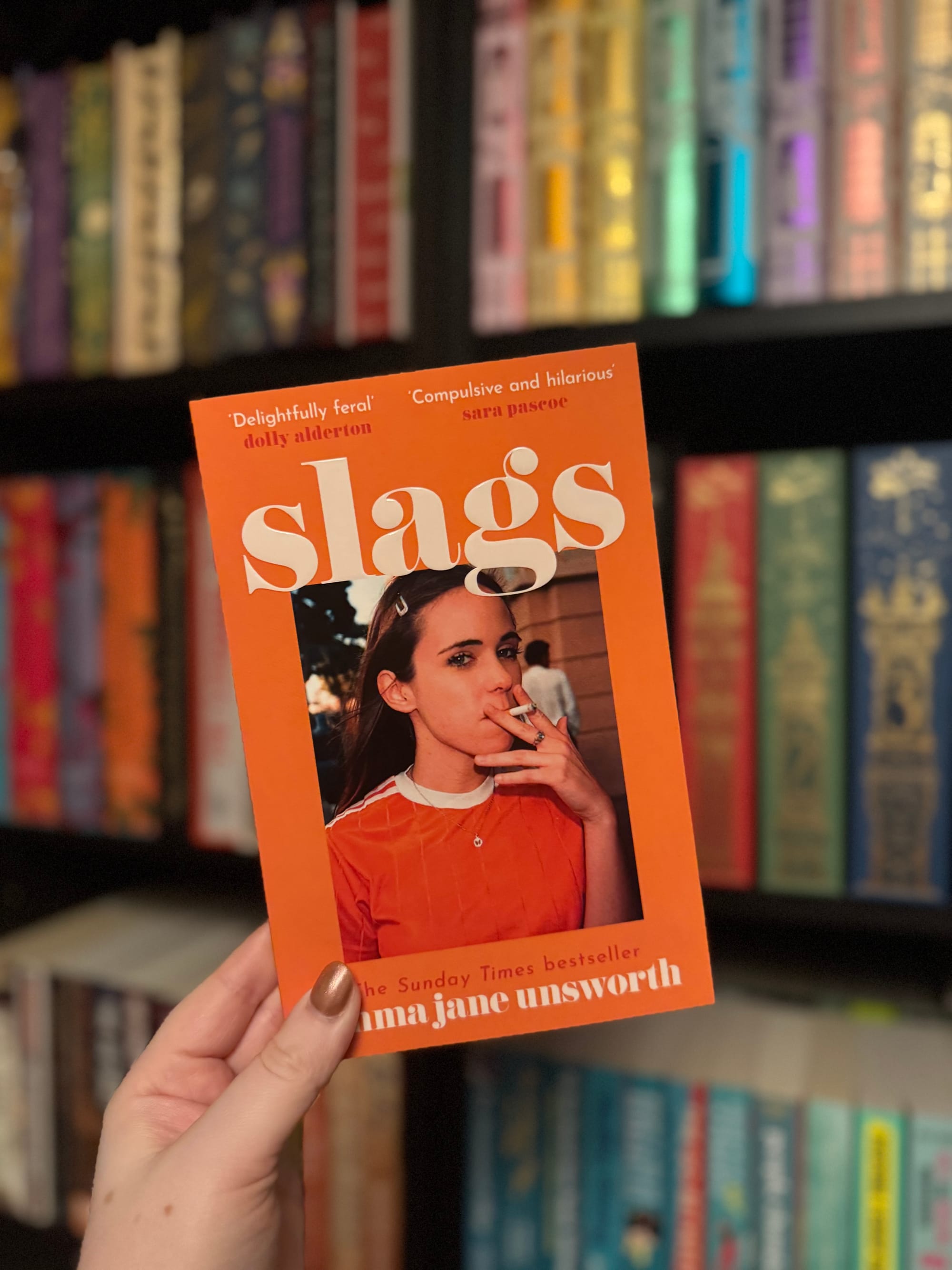 Hand holding book with orange cover titled "Slags." Woman on cover smokes. Background: colorful bookshelf. Moods: playful, rebellious.