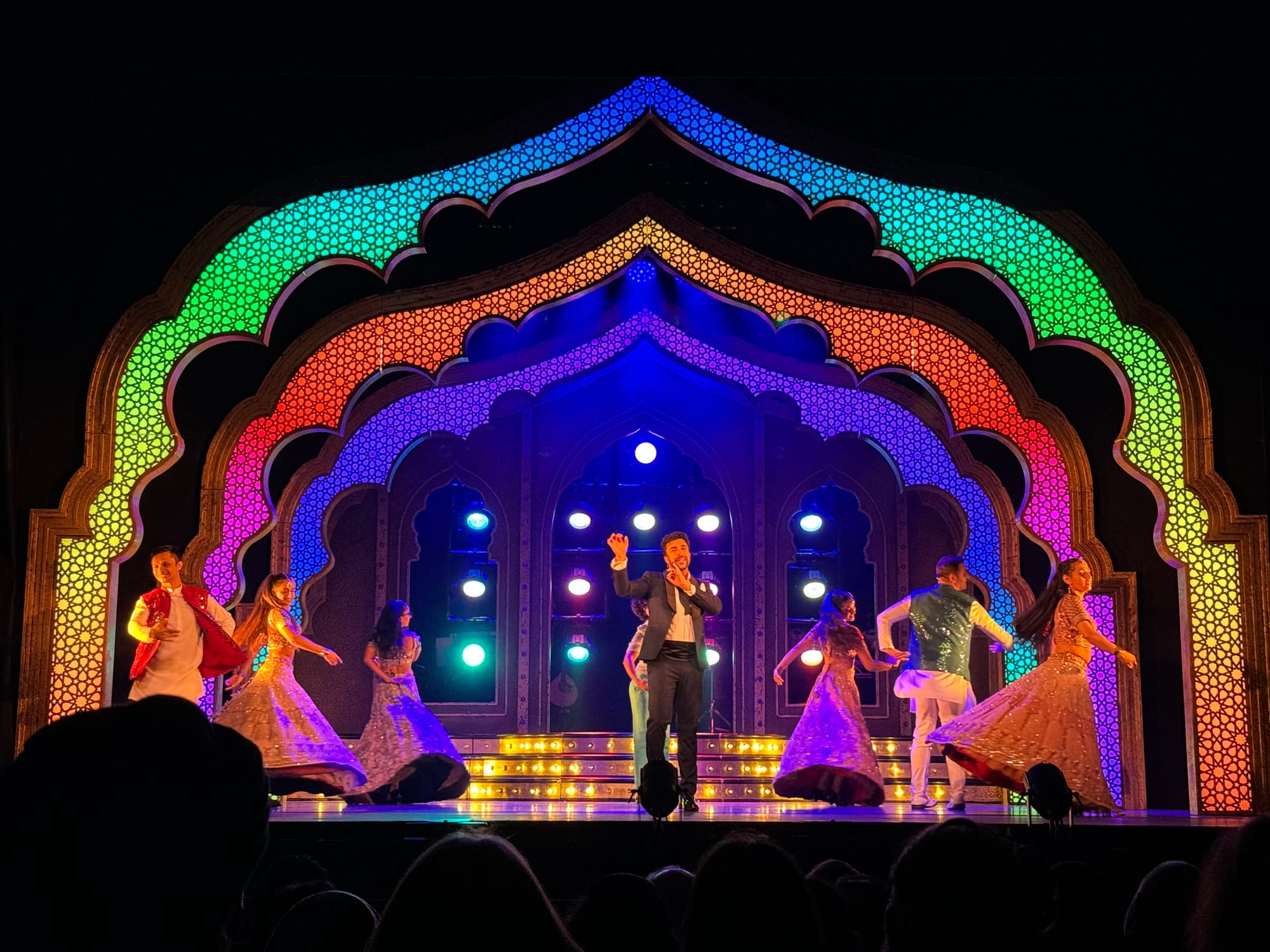 Performers in colorful traditional attire dance on a stage with a vibrant red arch backdrop, illuminated by warm spotlights, before an audience.