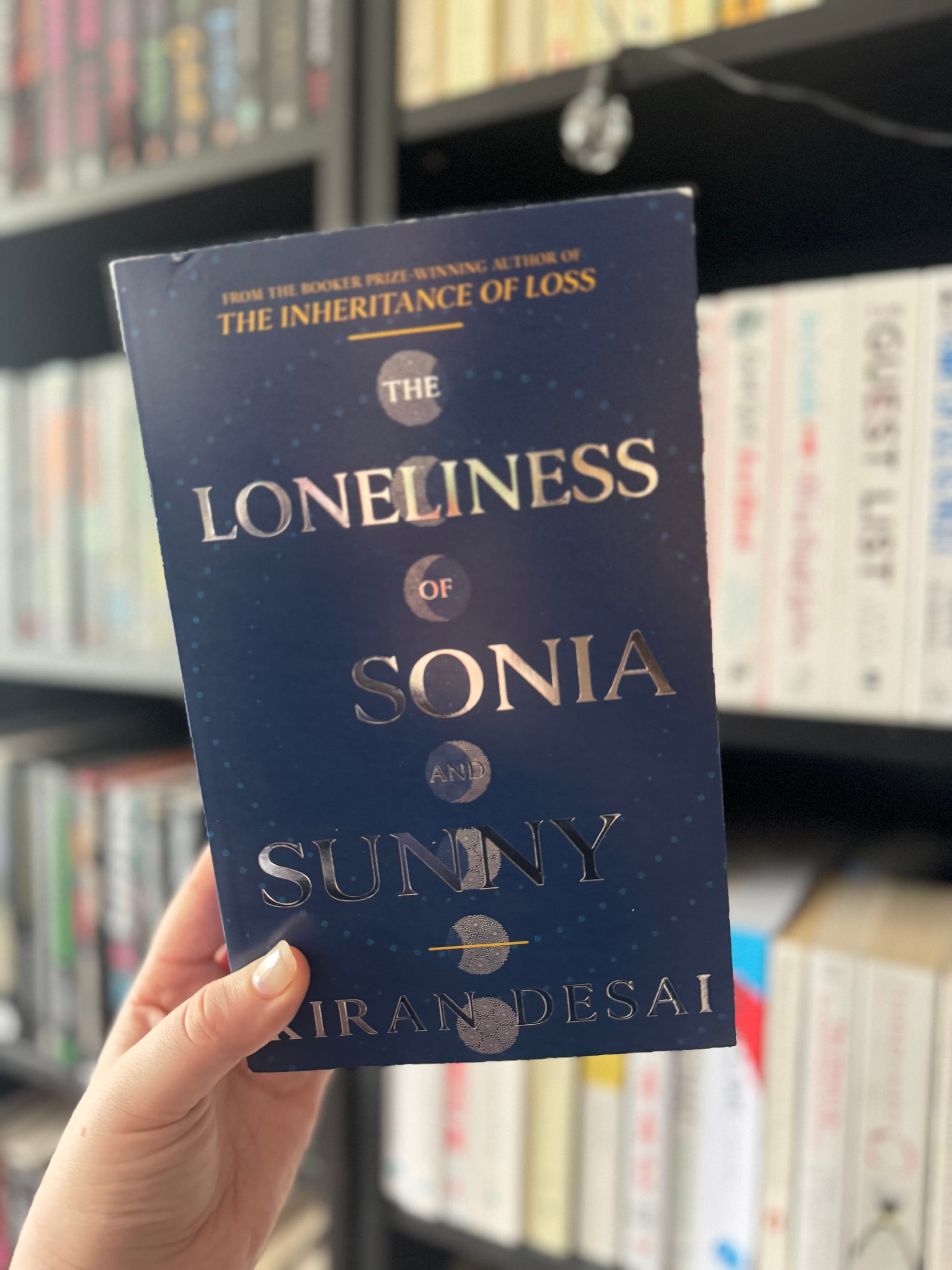 Hand holding a blue book, "The Loneliness of Sonia and Sunny" by Kiran Desai, in front of a blurred bookshelf background.