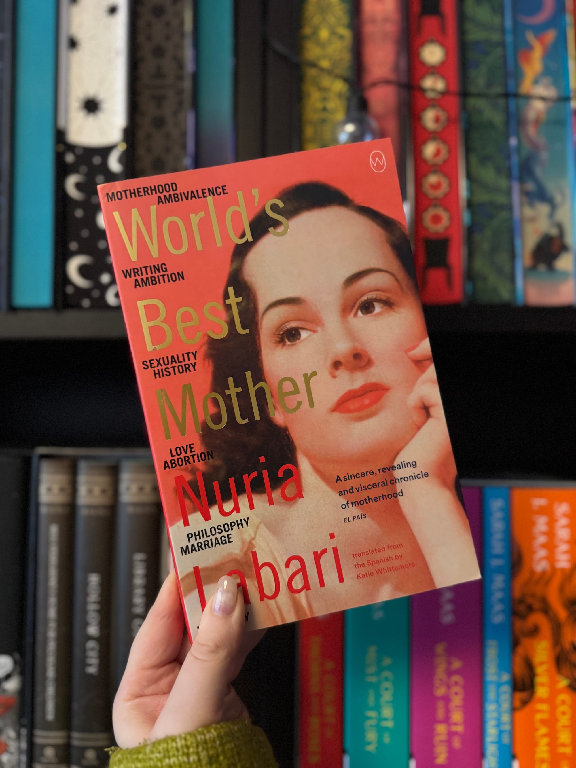 A hand holds the red cover of "World's Best Mother" by Nuria Labari, featuring a woman's face. Colorful books line the background shelves.