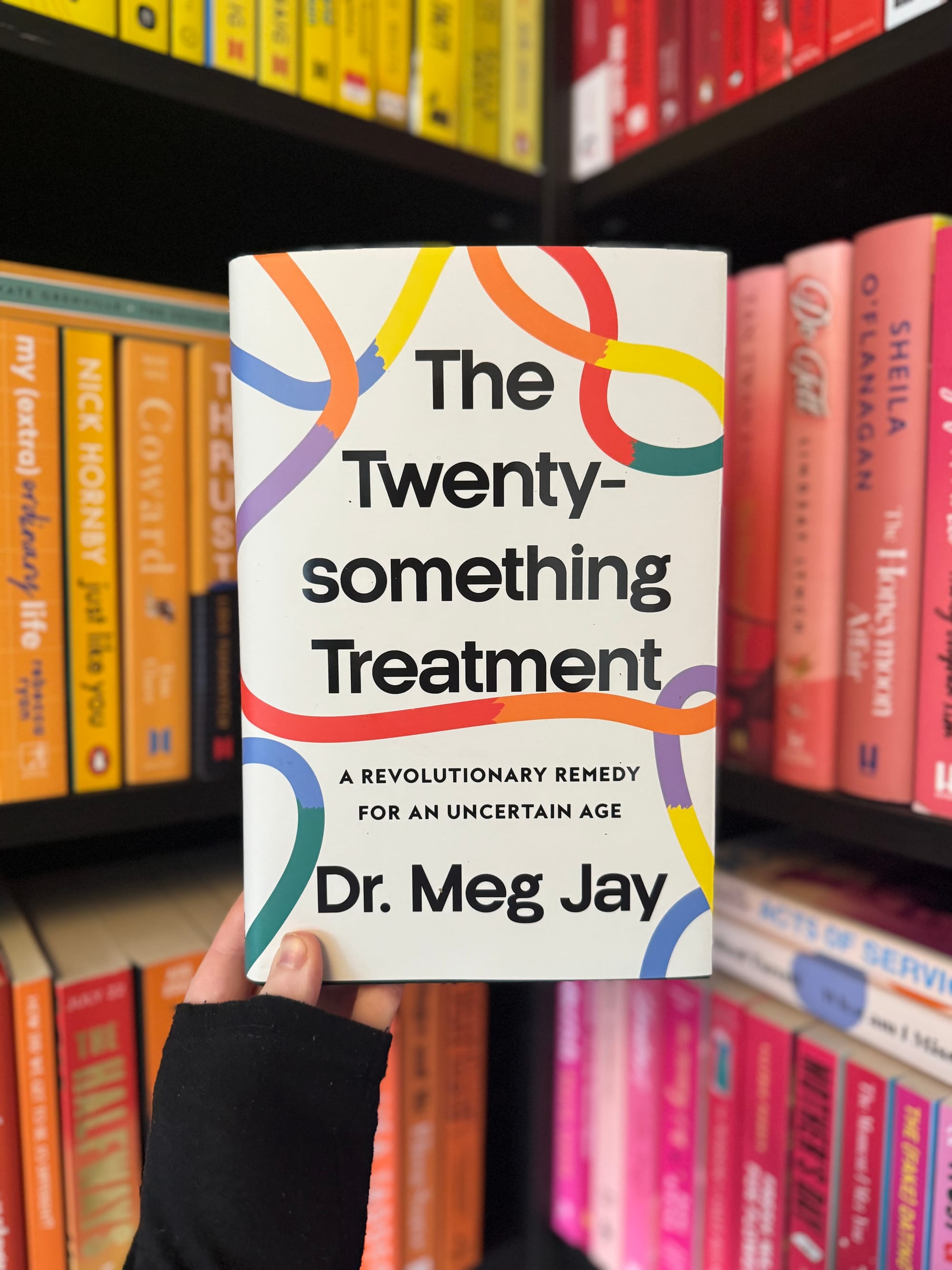 Hand holding "The Twenty-something Treatment" book by Dr. Meg Jay in front of colorful bookshelves with yellow and pink books.