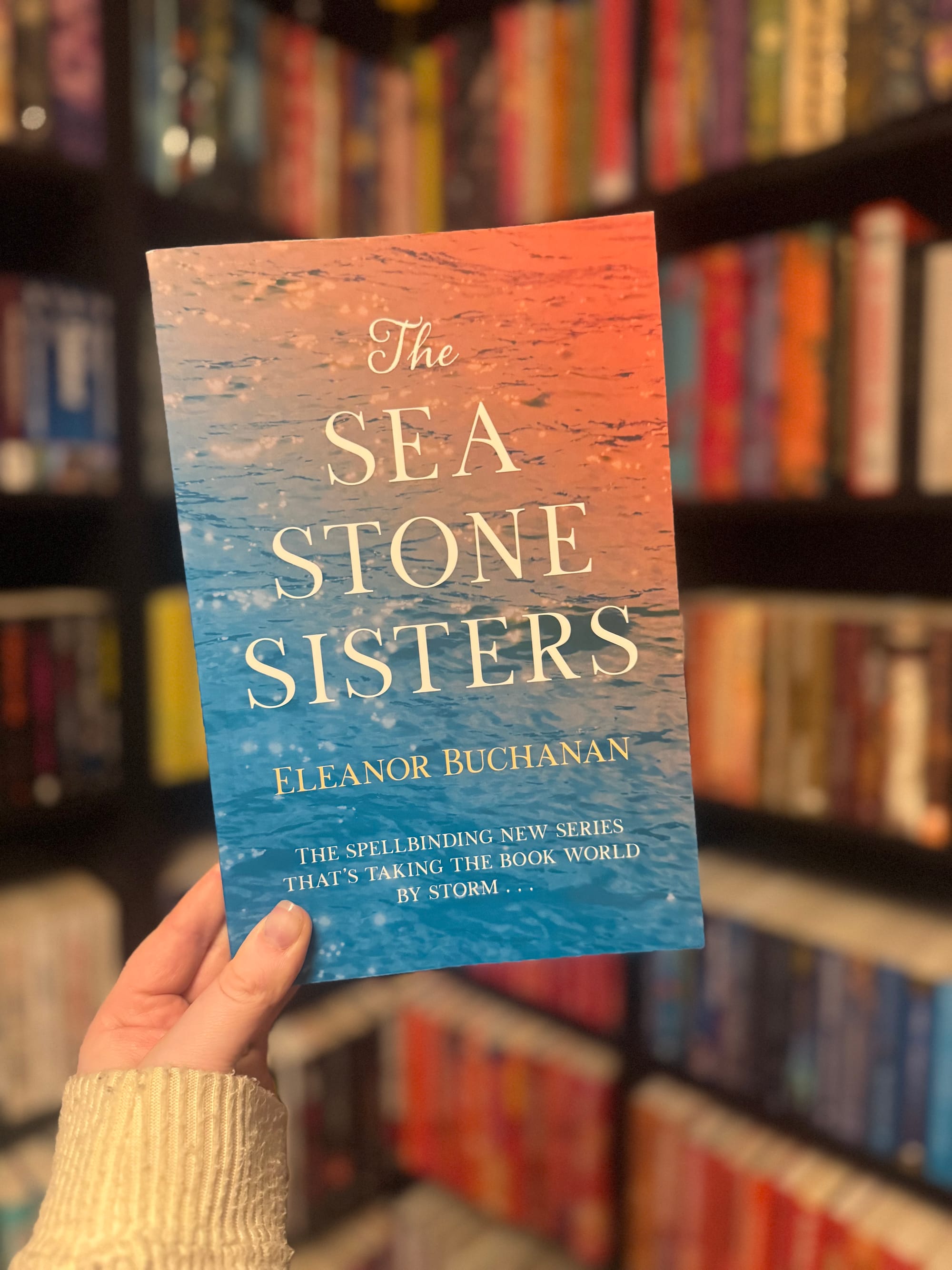 A hand holds a book titled "The Sea Stone Sisters" by Eleanor Buchanan against a blurred bookstore background, featuring colorful shelves.