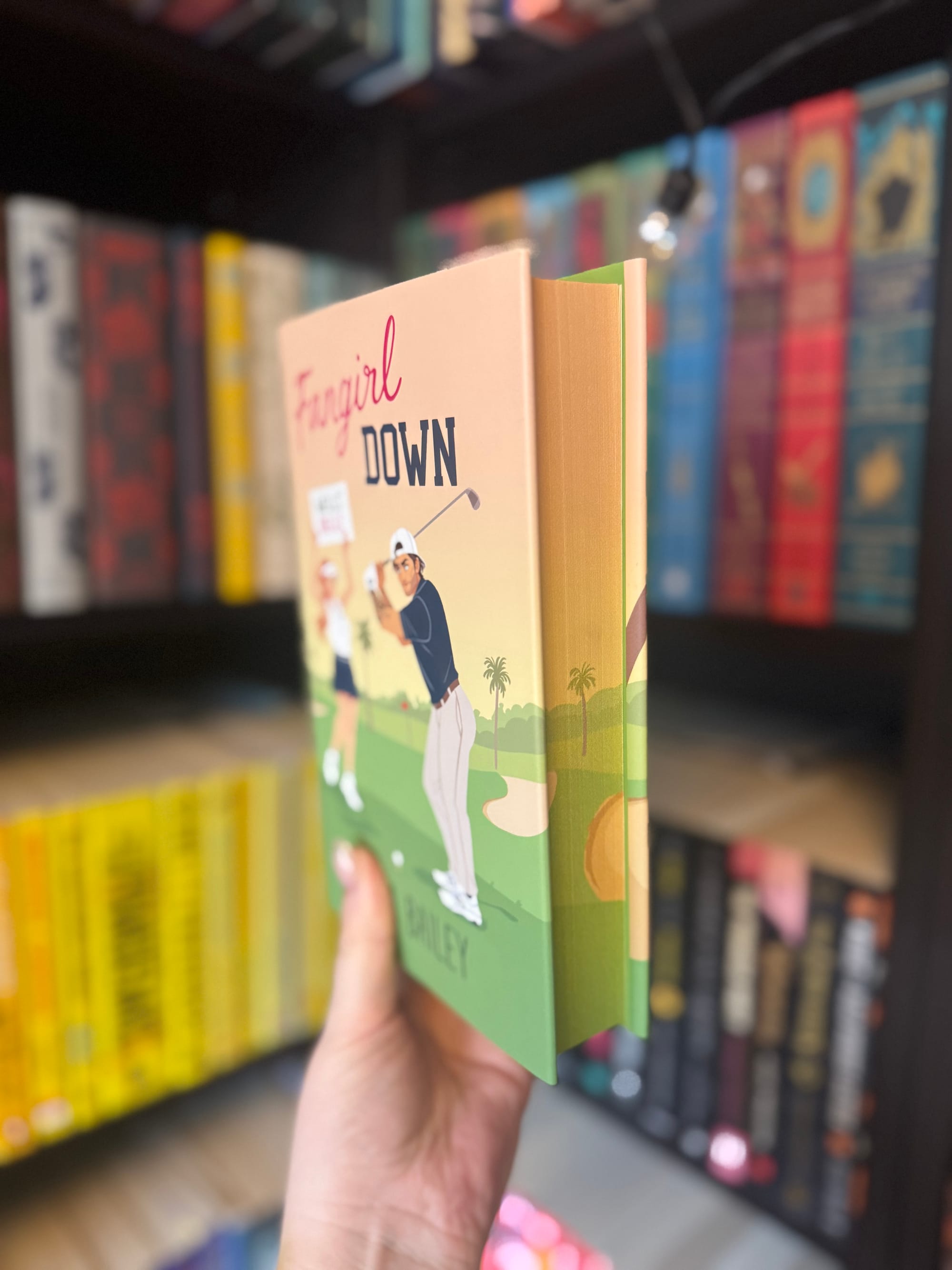 A hand holds a book titled "Fangirl Down" in front of colorful bookshelves. The cover shows a golfer on a course under blue skies.