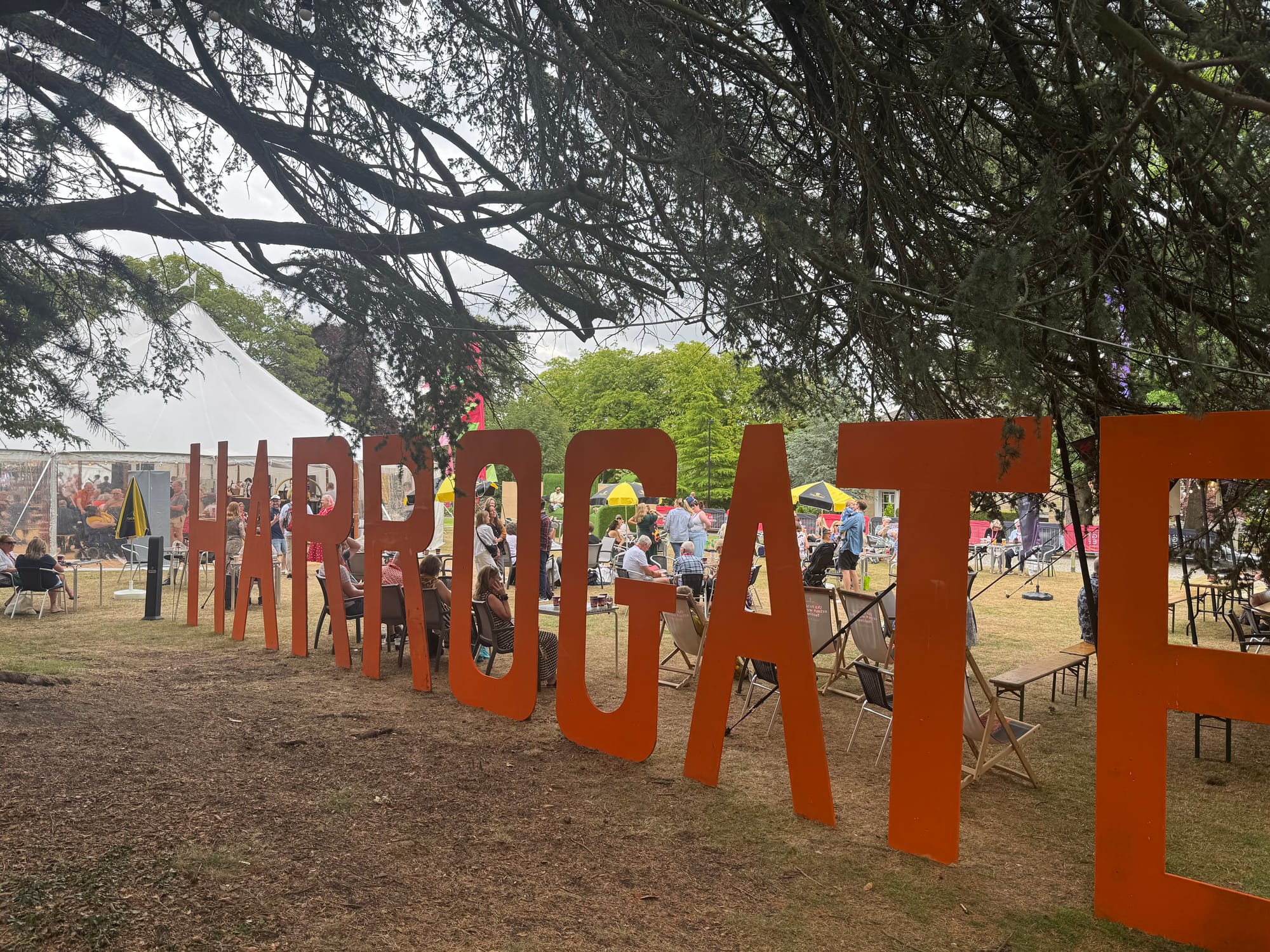 Outdoor view of the festival grounds with large orange letters spelling “HARROGATE” under a canopy of trees, with attendees relaxing at tables near tents and umbrellas.
