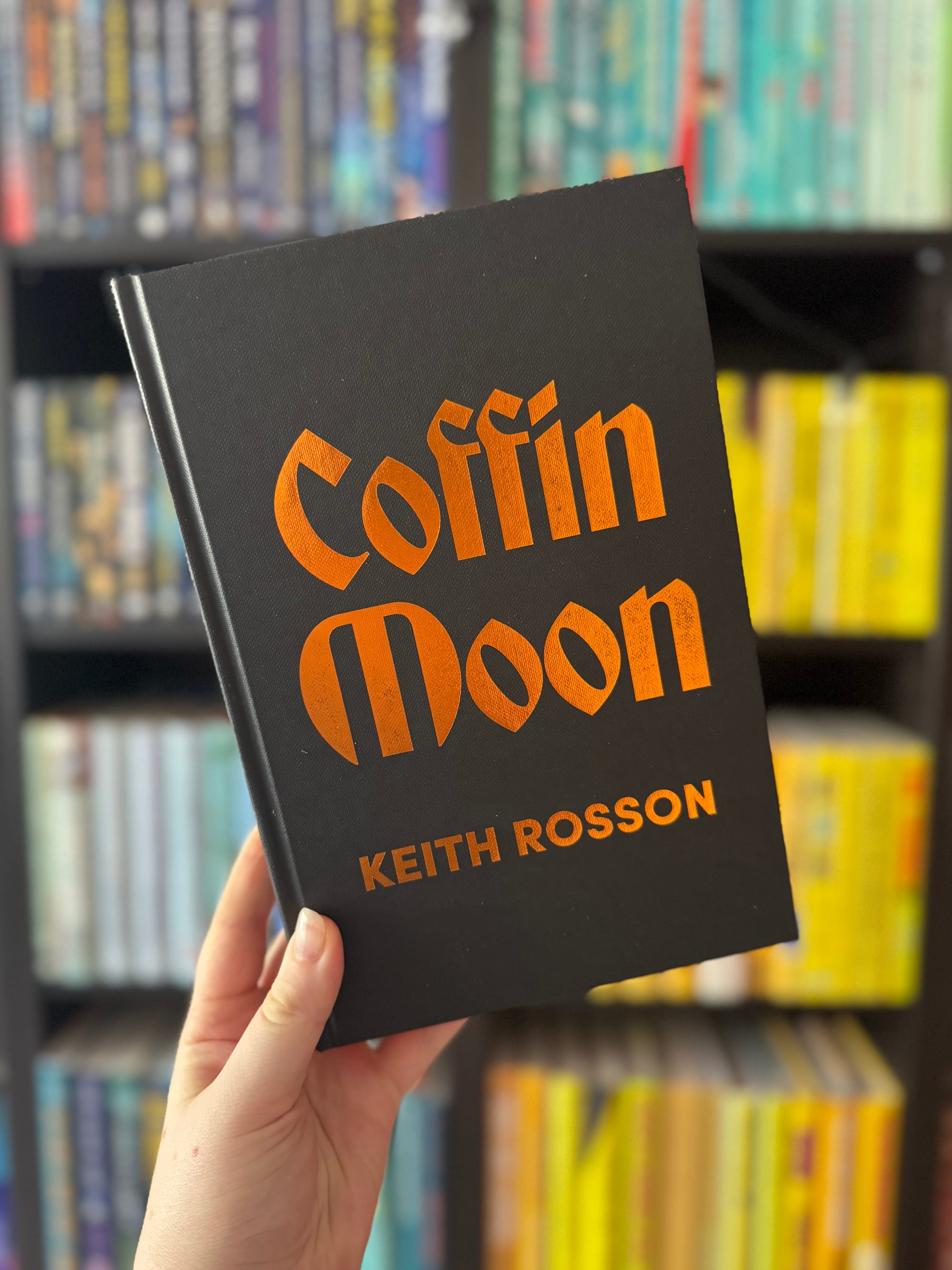 A hand holding the book 'Coffin Moon' by Keith Rosson. The book has a black cover with the title and author’s name embossed in bright orange text. In the background, there are bookshelves filled with colourful books arranged by spine colour.