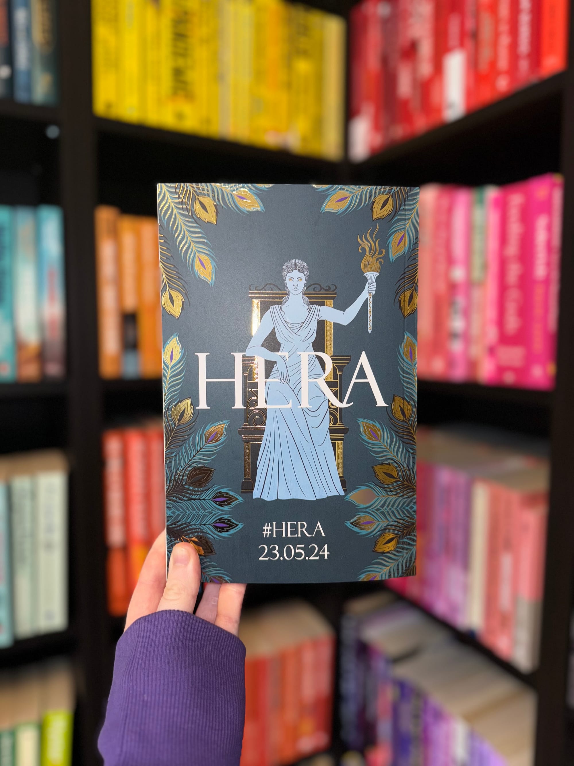 Hand holds a book titled "HERA" featuring a figure with a torch, against a colorful bookshelf. Text: "#HERA 23.05.24". Purple sleeve visible.