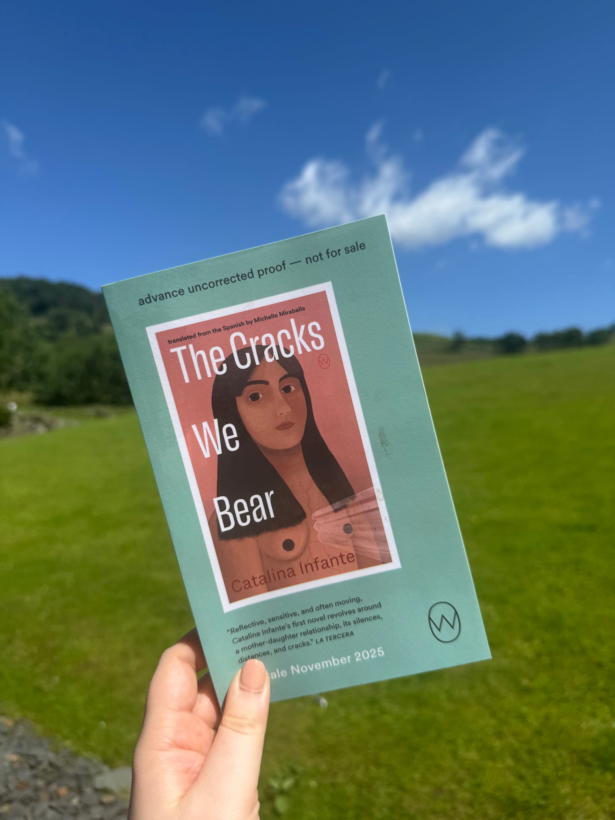 Hand holding a book titled "The Cracks We Bear" by Catalina Infante, against a grassy field and blue sky. Text includes "advance uncorrected proof."