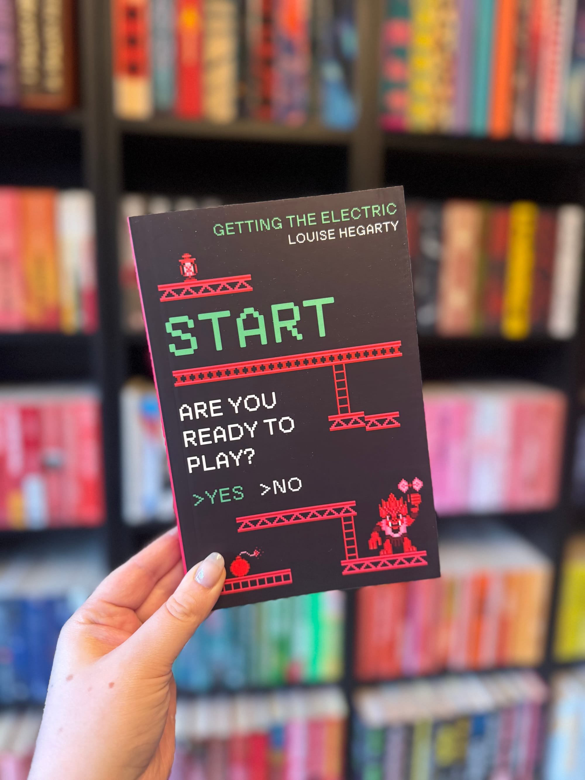 Hand holds a book titled "Getting The Electric" by Louise Hegarty, mimicking a video game start screen. Blurred colorful bookshelf background.