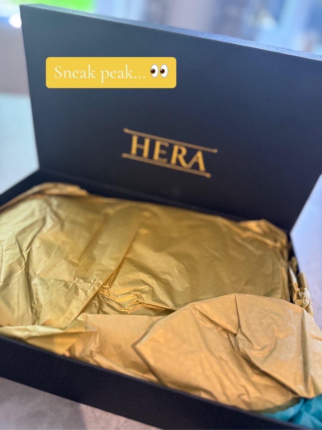 Open black box labeled "HERA" with crumpled gold tissue paper inside. Text overlay reads "Sneak peak..." with eye emoji.