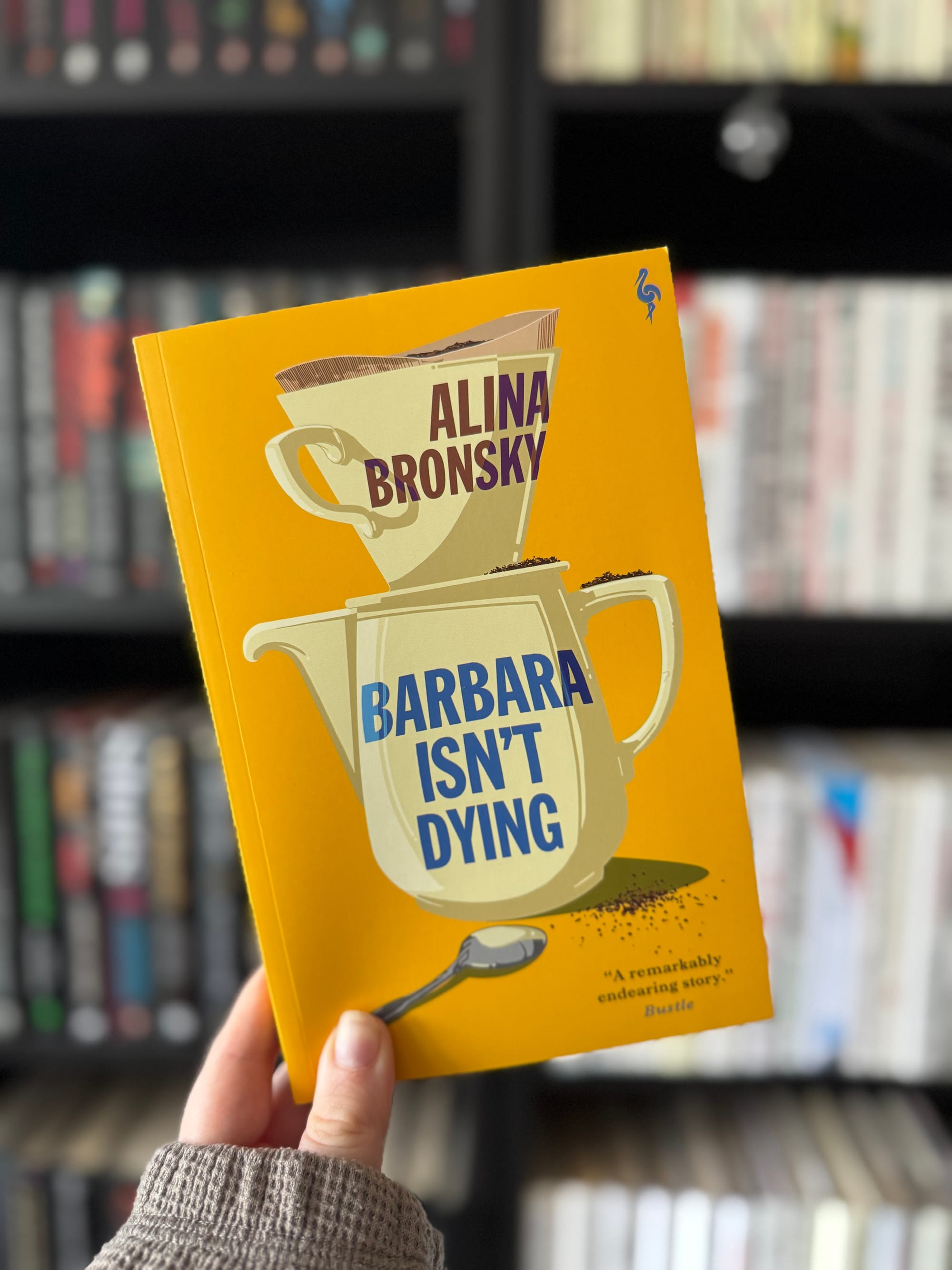 A hand holds a yellow book titled "Barbara Isn't Dying" by Alina Bronsky. Background shows a blurred bookshelf, setting a cozy library vibe.