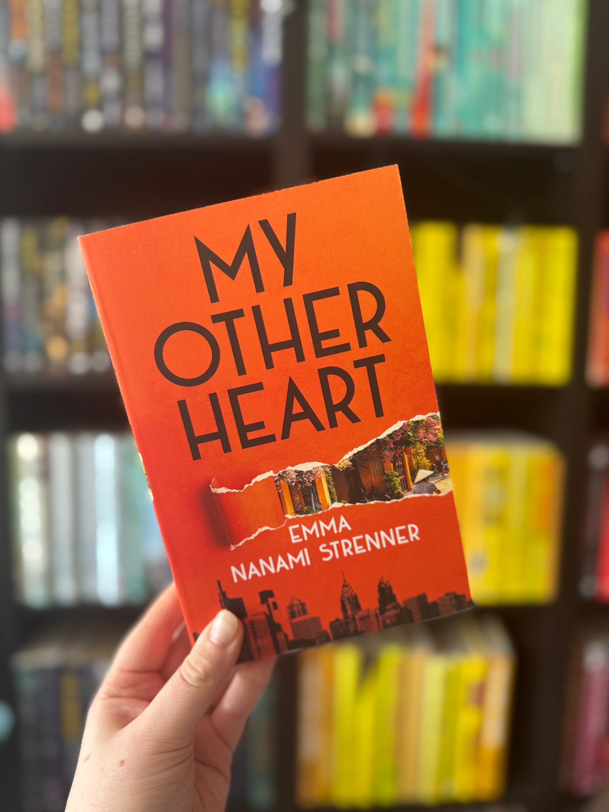 Hand holds an orange book titled "My Other Heart" by Emma Nanami Strenner, with a blurred bookshelf background filled with colorful books.