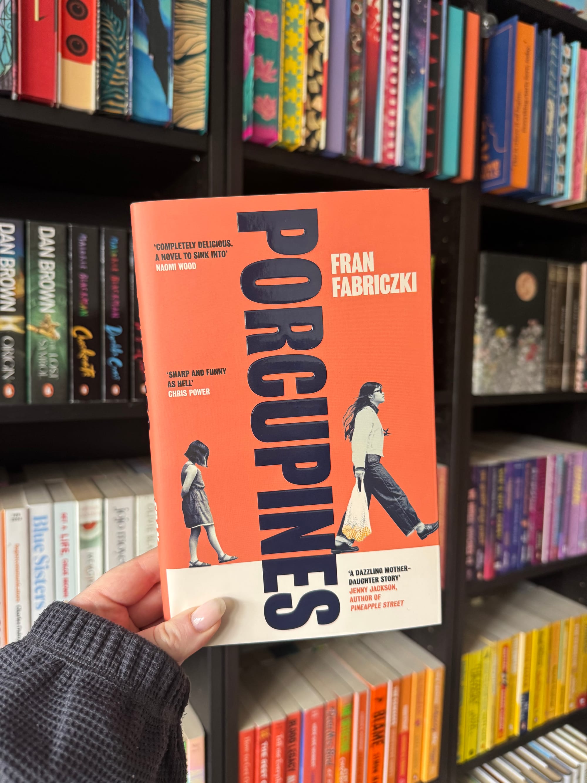 Hand holding "Porcupines" book by Fran Fabriczki in a colorful bookstore. The cover is orange with an illustration of a woman and girl.