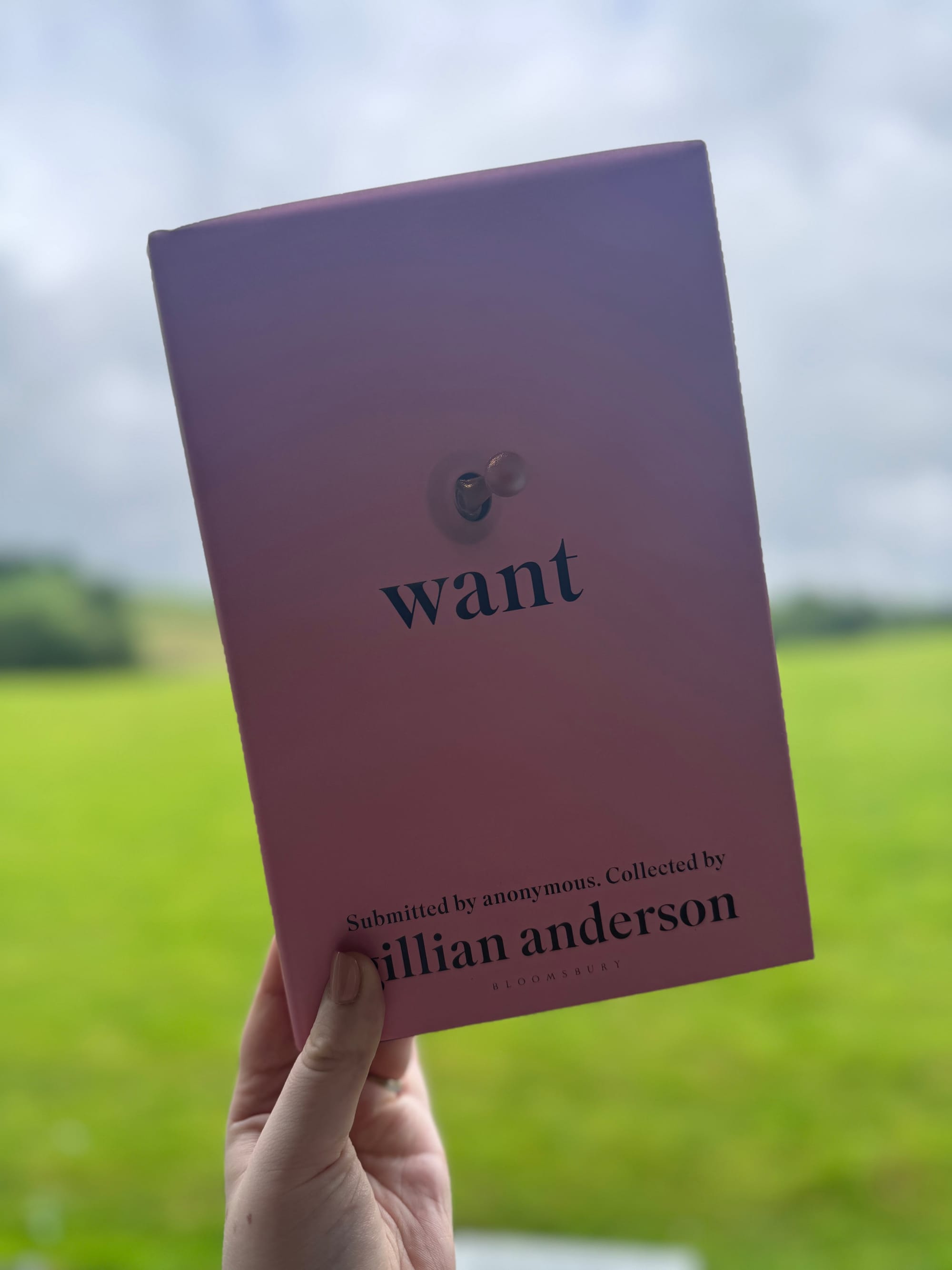 A hand holds a pink book titled "Want" by Gillian Anderson against a blurred green field and cloudy sky, conveying a calm outdoor setting.