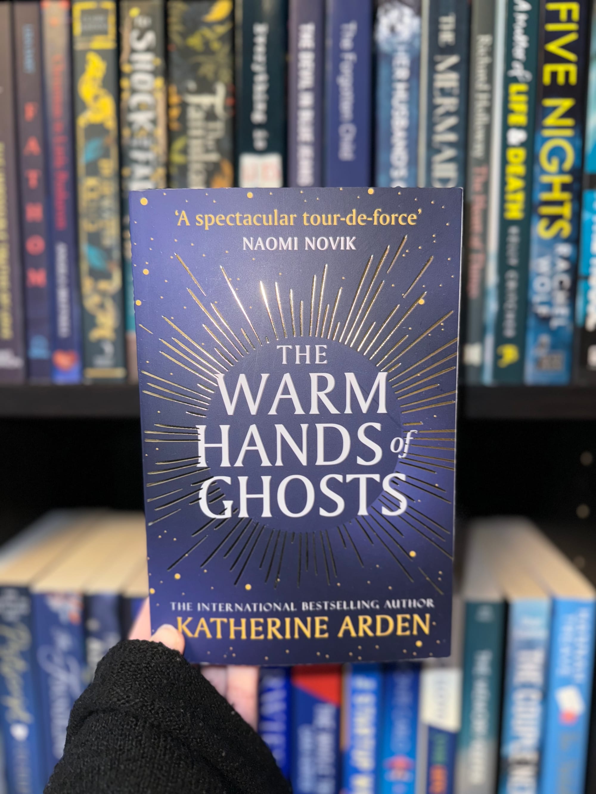 Hand holds "The Warm Hands of Ghosts" book by Katherine Arden. Blue cover with gold design. Shelf with other books in background.