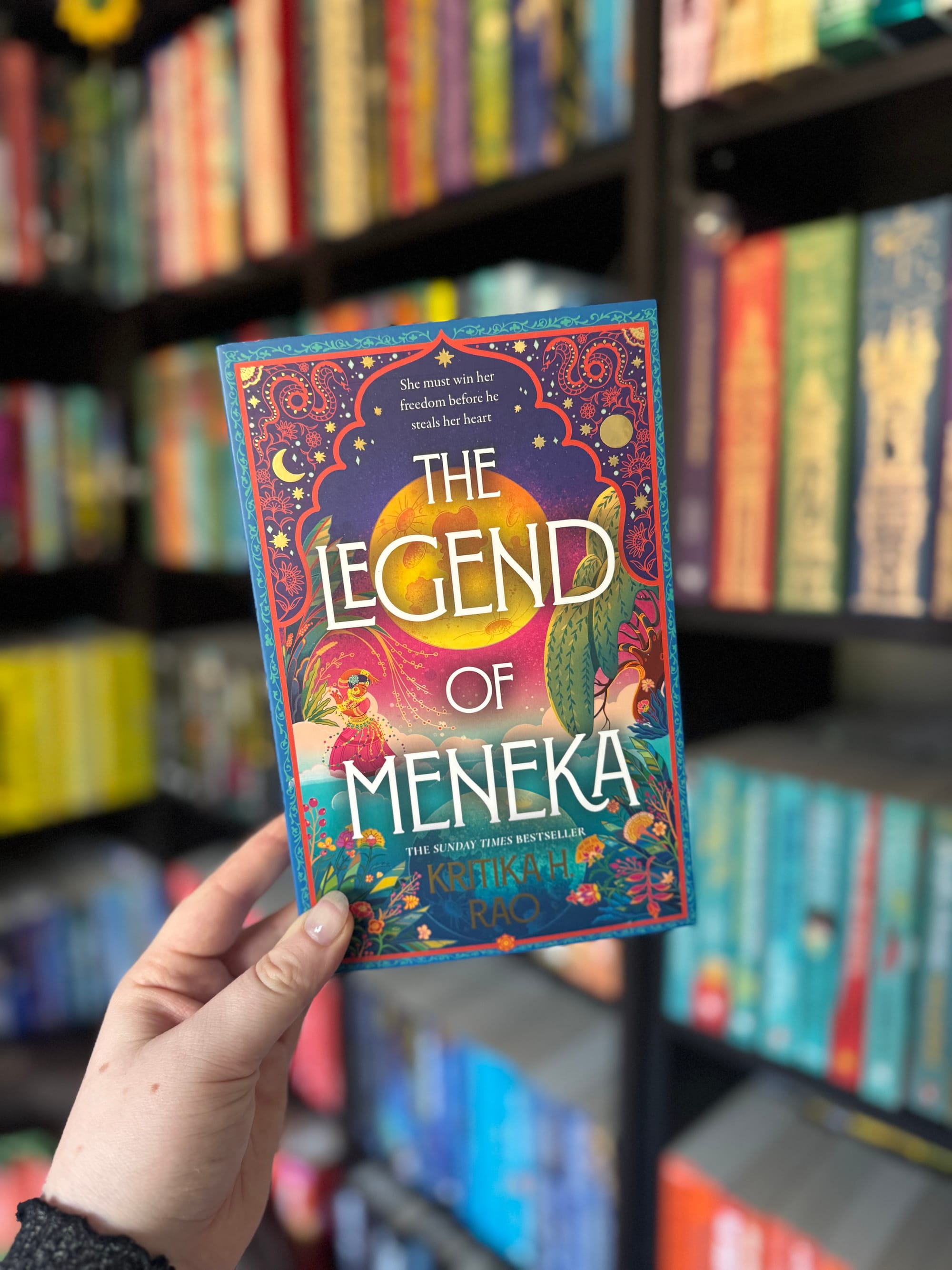 Hand holding colorful book "The Legend of Meneka" in front of a vibrant bookshelf. Elegant cover art features intricate patterns.