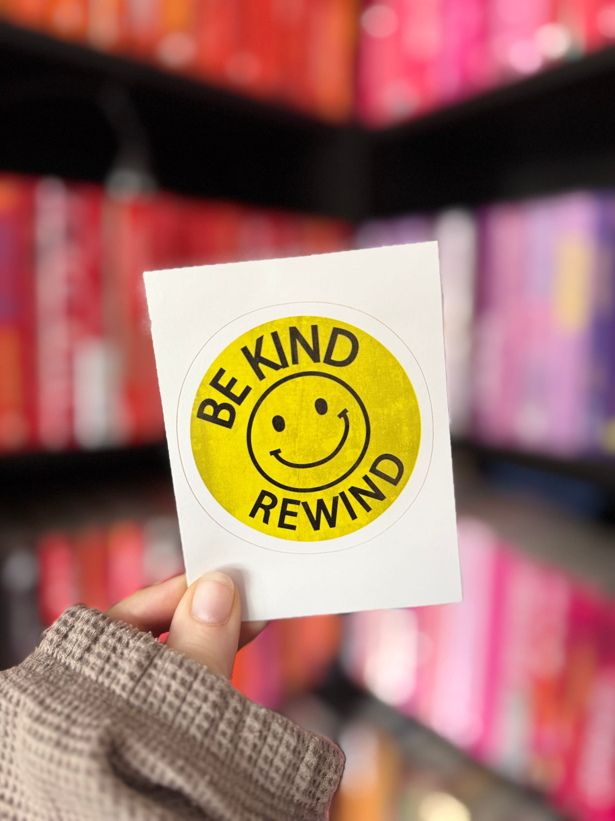 A hand holds a retro-style yellow sticker with a smiley face and the text "BE KIND REWIND" in black letters. The background features a colourful bookshelf arranged by spine colour, with red, pink, and purple tones visible.