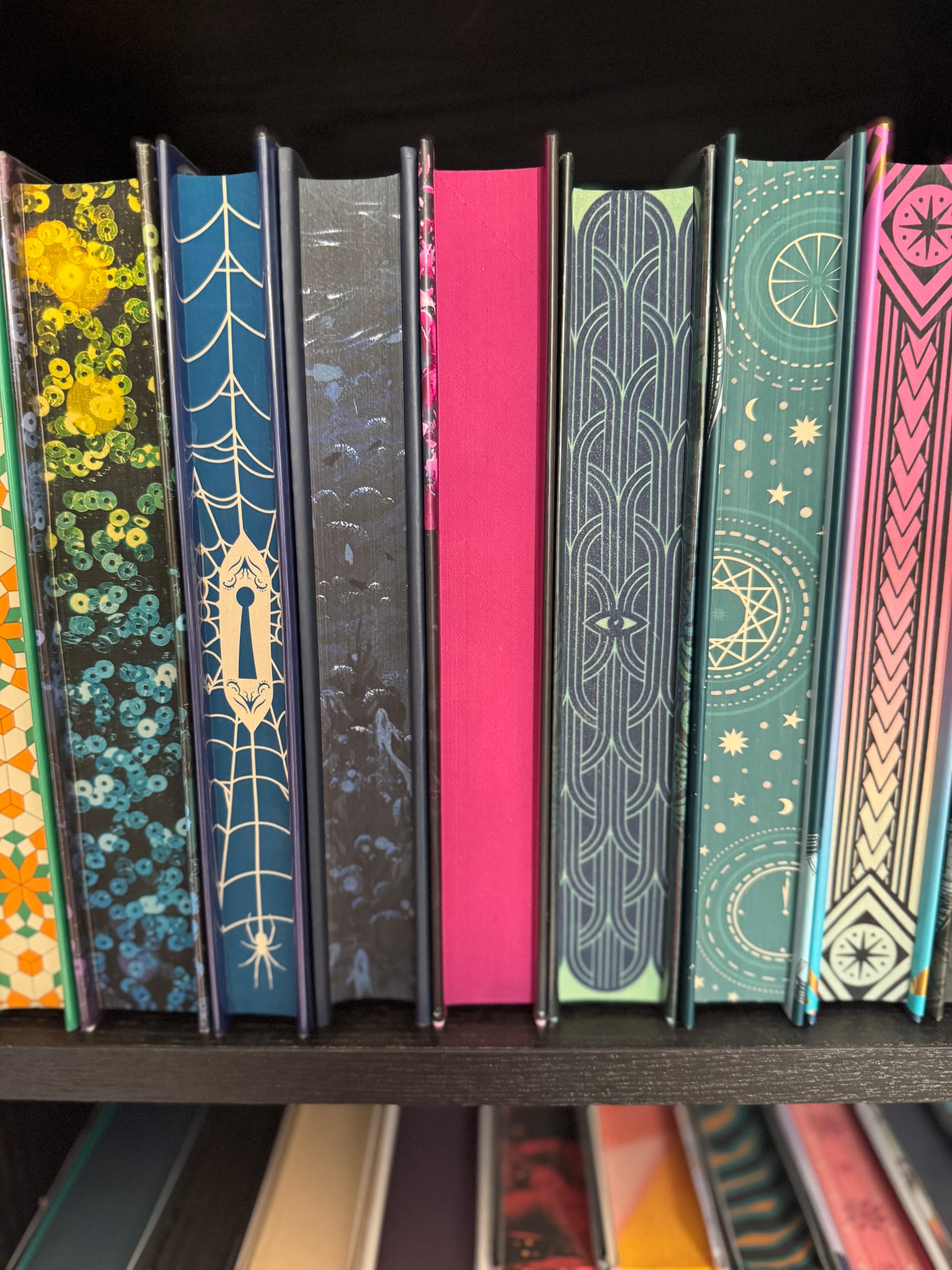 Colorful books with patterned spines stand on a black shelf. Designs include stars, webs, and geometric shapes, creating a vibrant display.