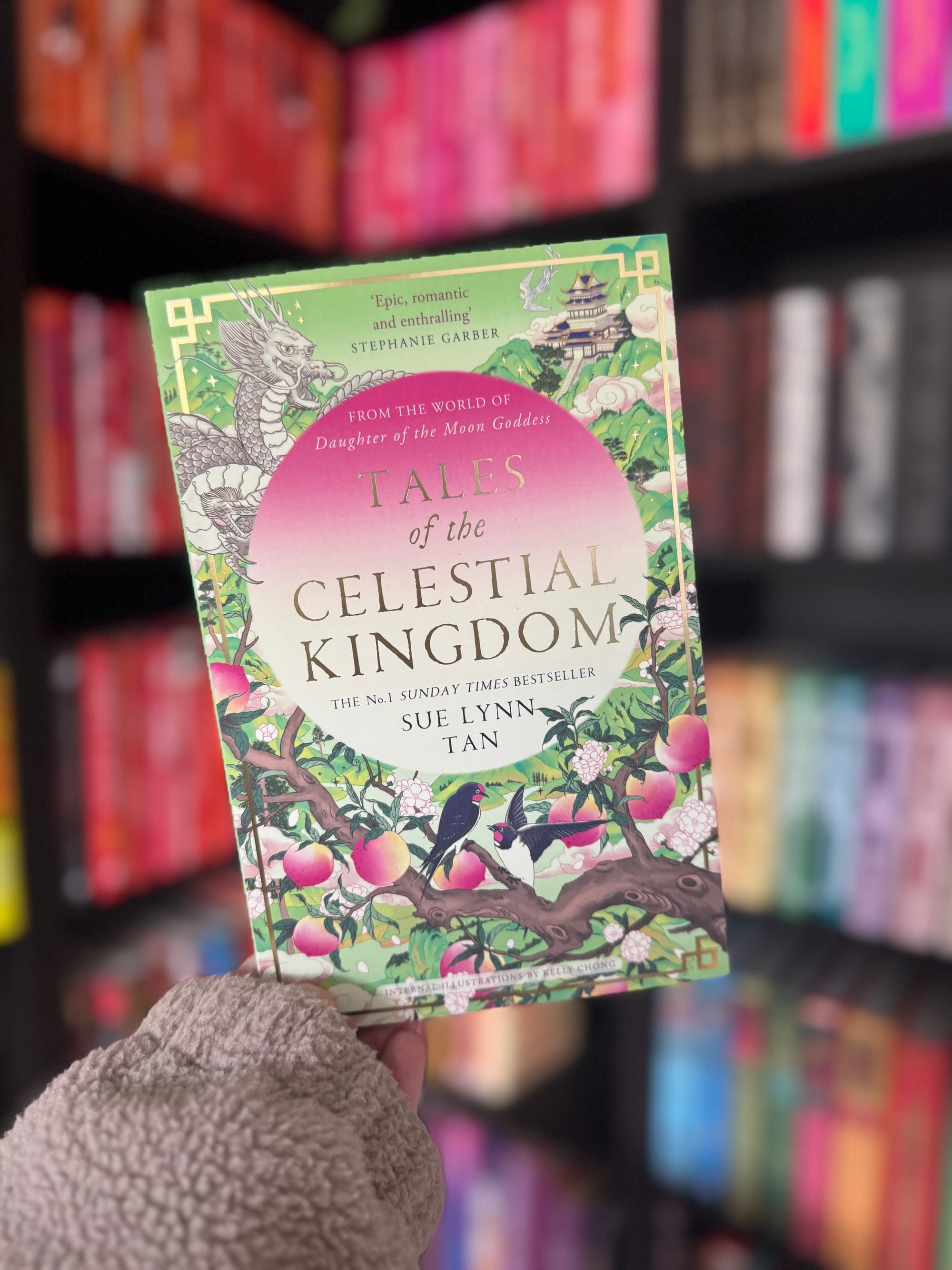 A hand holds "Tales of the Celestial Kingdom" book in a library. The cover features a dragon, flowers, and birds. Background shows colorful books.