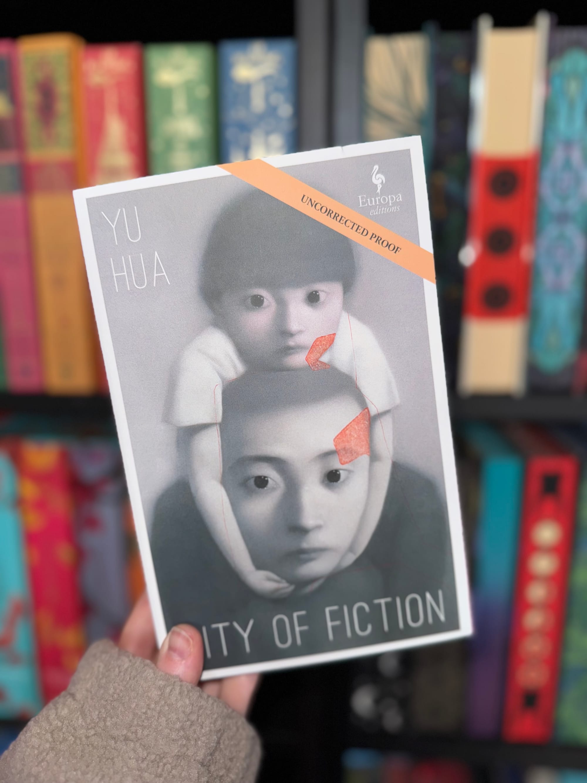 Hand holds a book titled "City of Fiction" by Yu Hua, labeled "Uncorrected Proof," against a colorful bookshelf background.