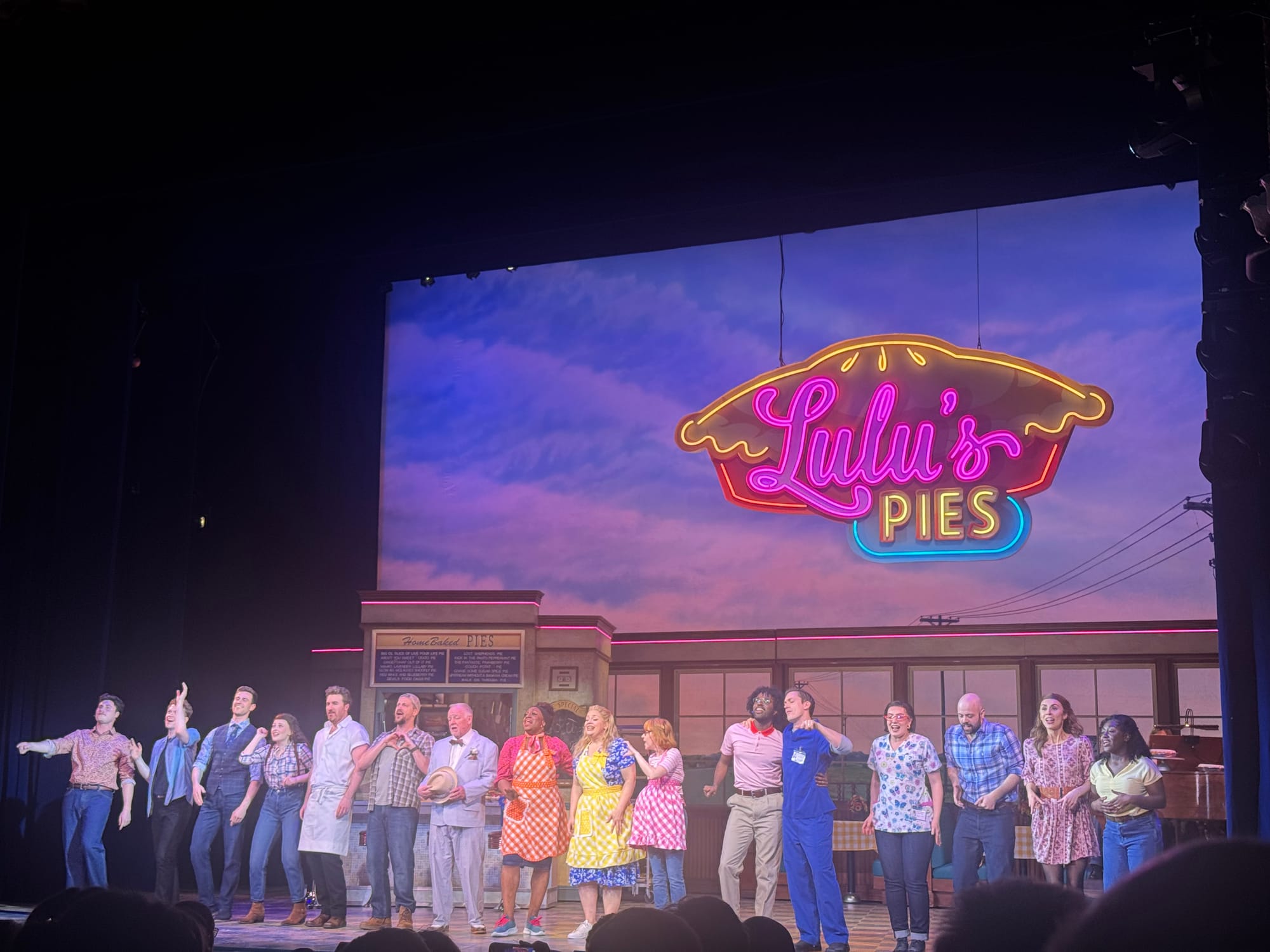 Full cast of Waitress the Musical taking a bow on stage in front of Lulu’s Pies diner set with neon sign backdrop
