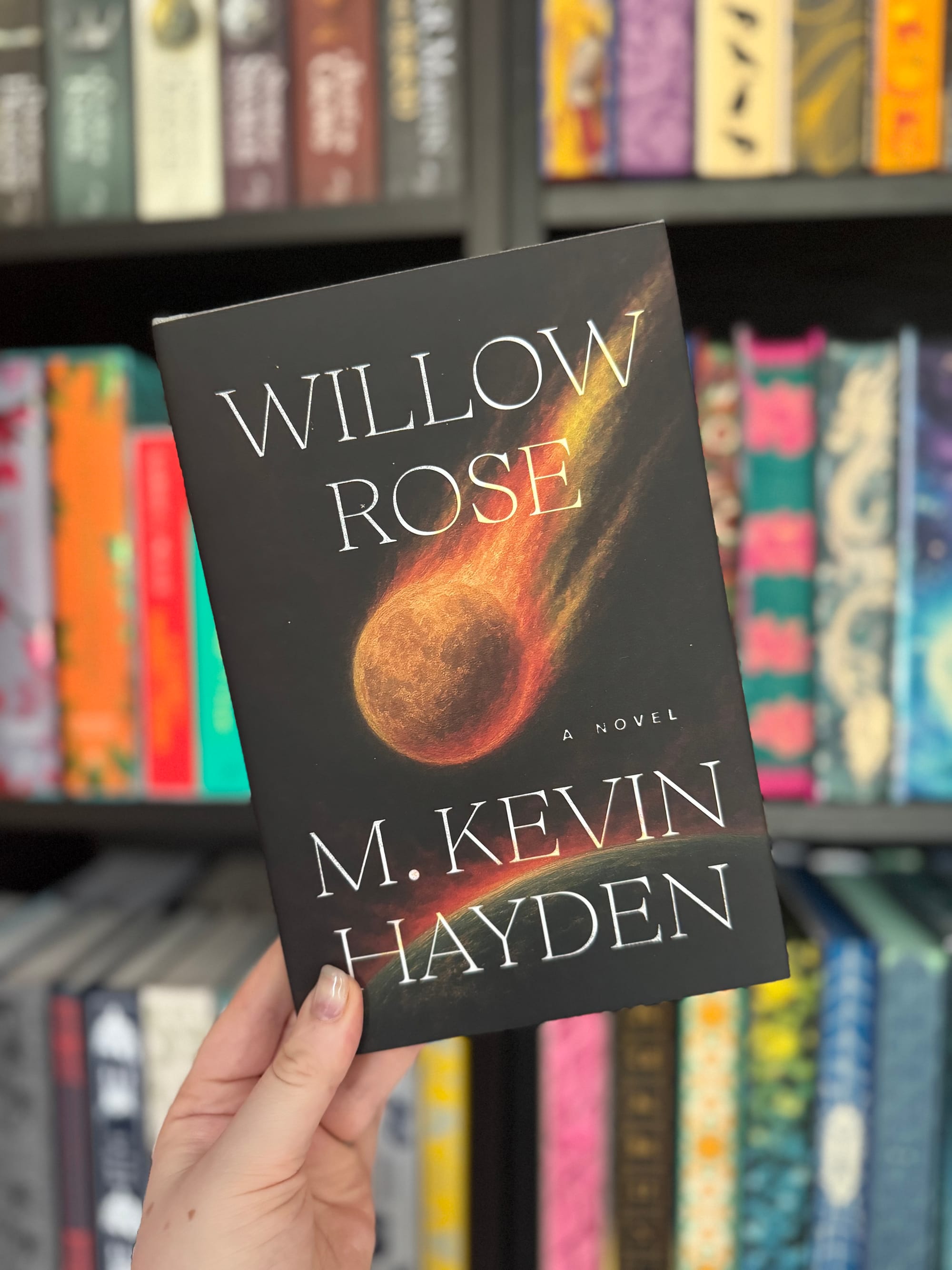 A hand holds the book "Willow Rose" by M. Kevin Hayden with a fiery cover. Bookshelf with colorful books in the blurred background.