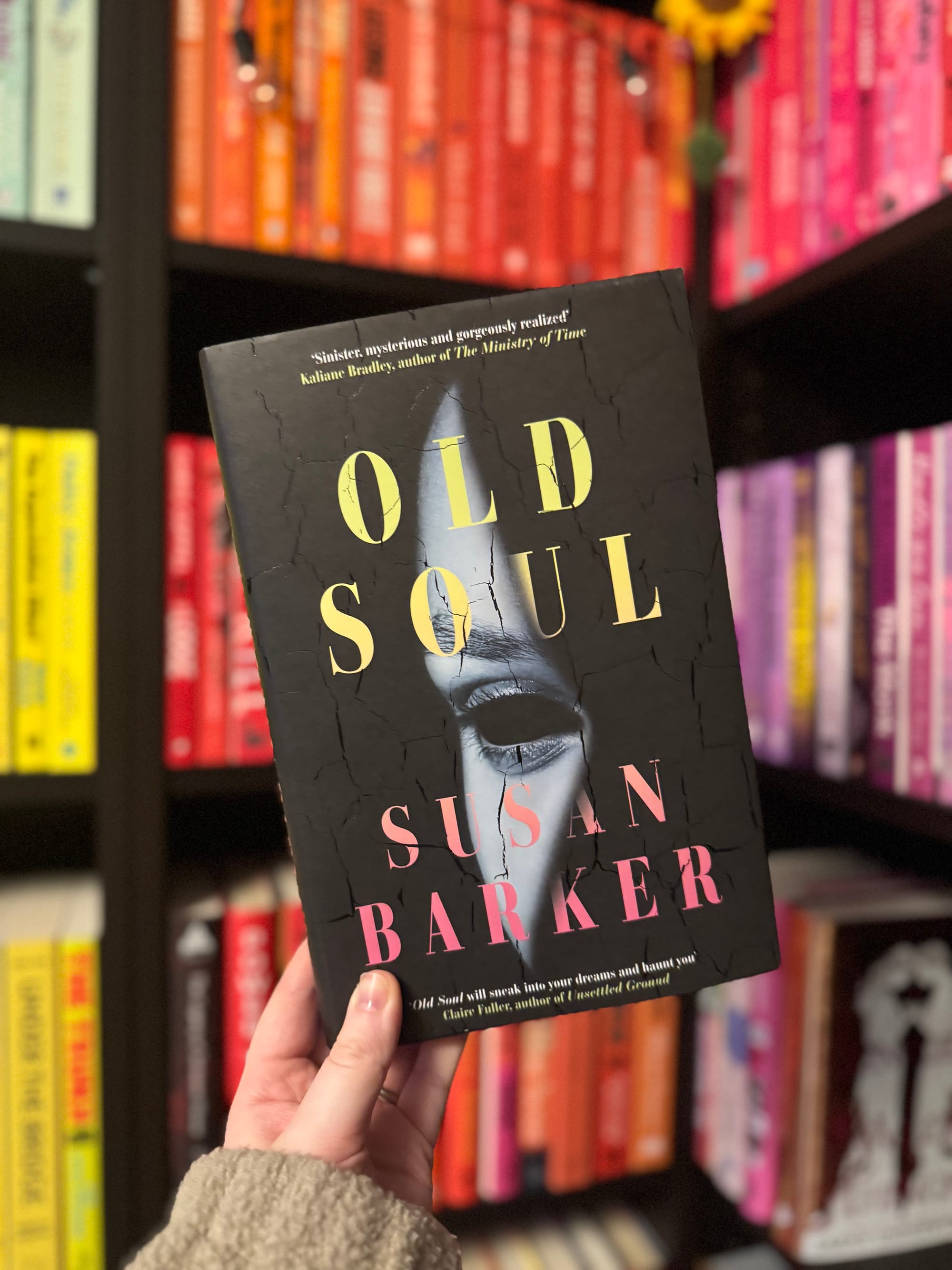 Hand holding the book "Old Soul" by Susan Barker, with cracked cover design. Background shows colorful bookshelf with neatly arranged books.