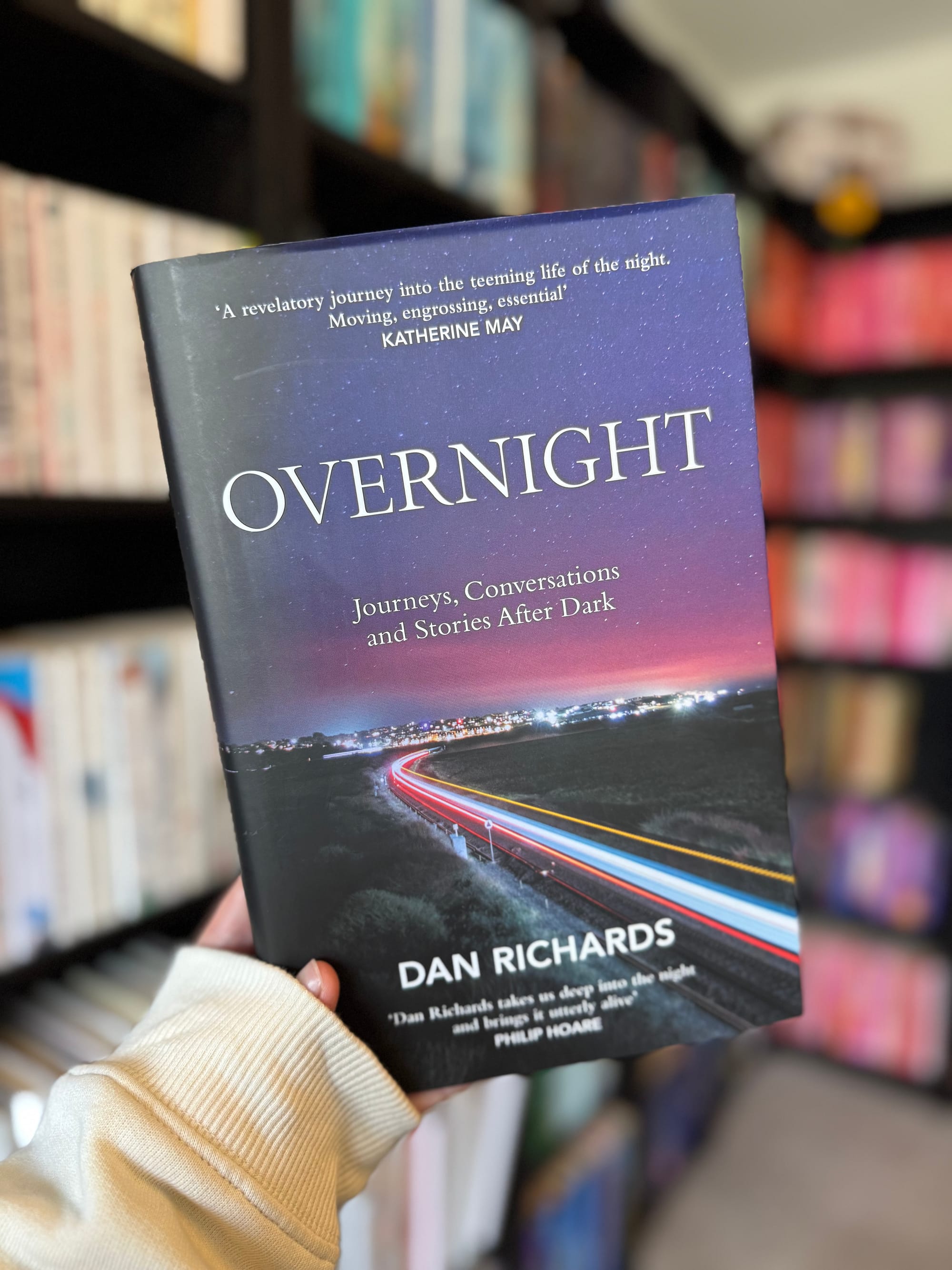 Person holds "Overnight" book by Dan Richards in a library. Cover shows a colorful night scene with light trails. Shelves of books in background.
