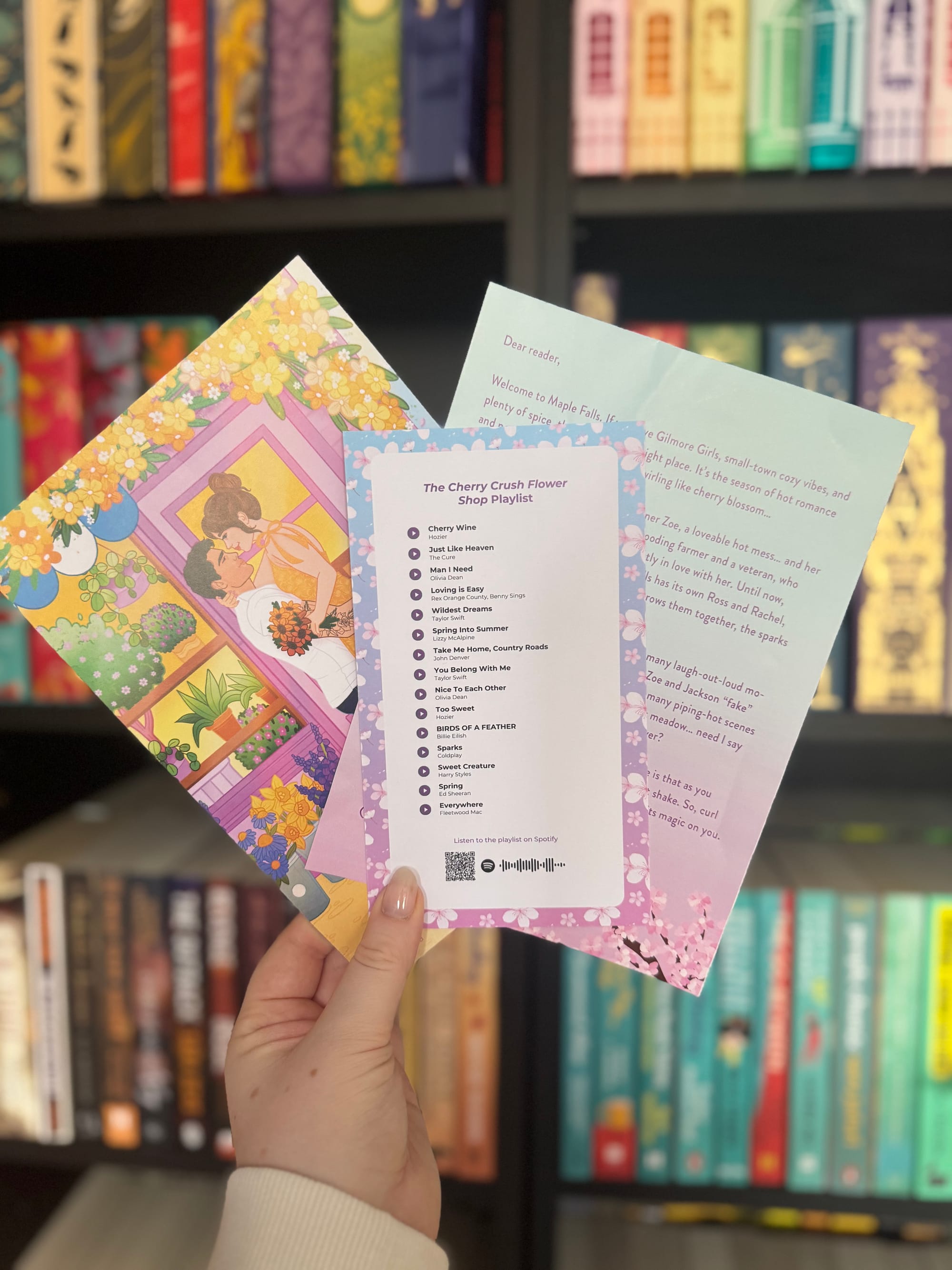 A hand holding three pastel themed book inserts in front of a bookshelf, including an illustrated card, a cherry blossom themed playlist card titled “The Cherry Crush Flower Shop Playlist,” and a welcome letter.