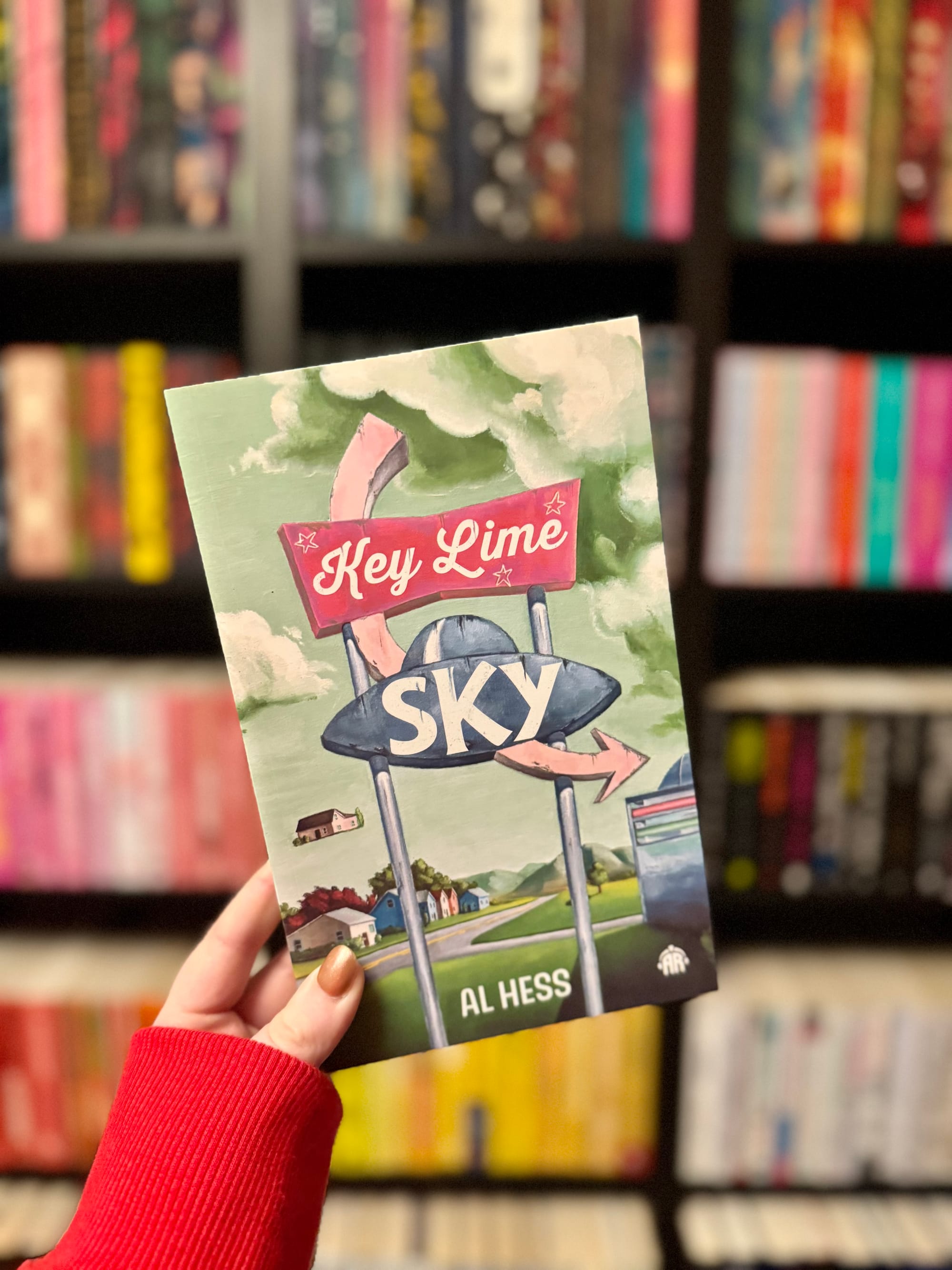 Hand holding a book titled "Key Lime Sky" by Al Hess, featuring a retro road sign illustration, in front of a colorful bookshelf background.