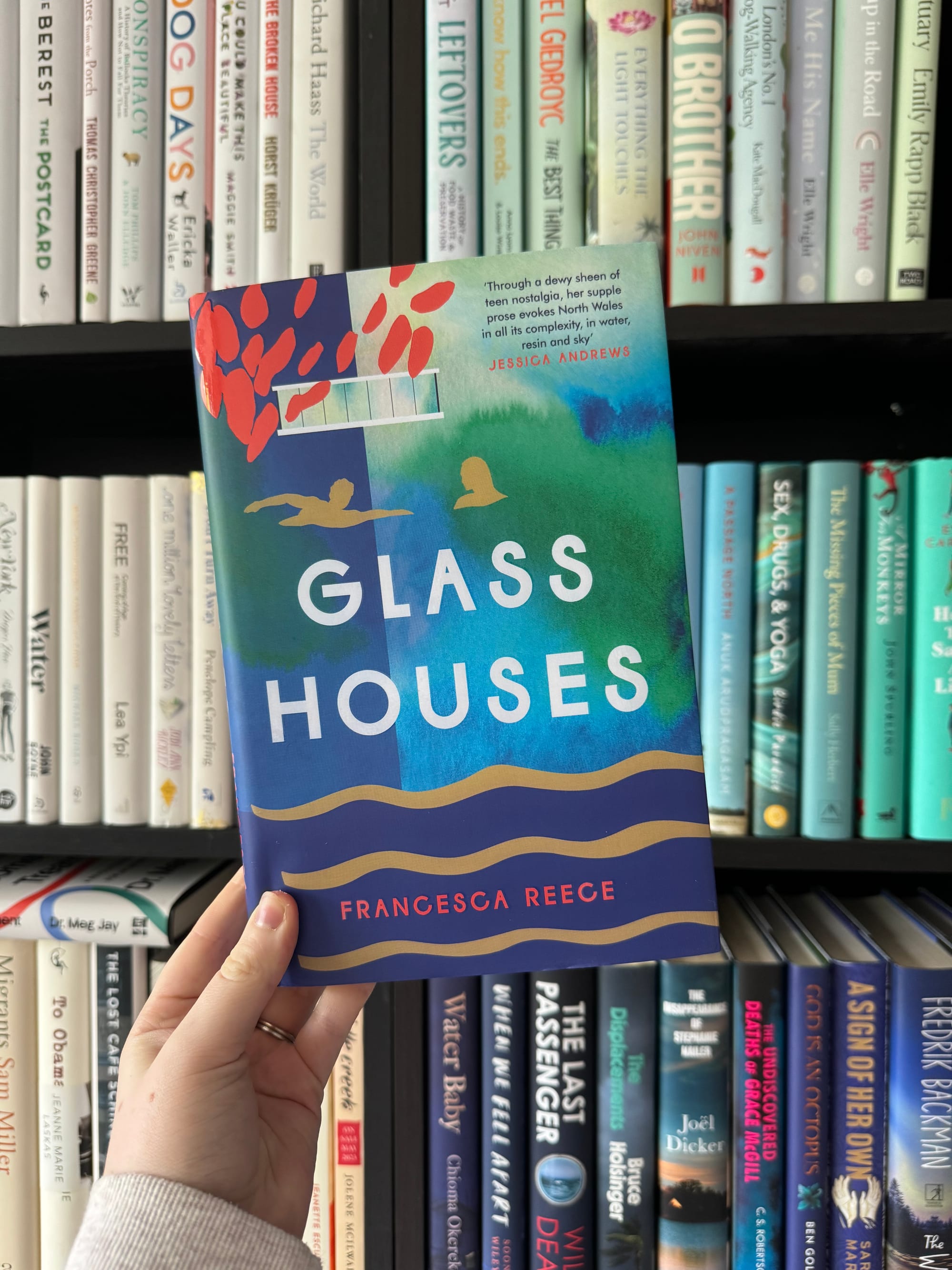 A hand holds the book "Glass Houses" by Francesca Reece against a bookshelf. The cover features abstract designs in red, blue, and green.
