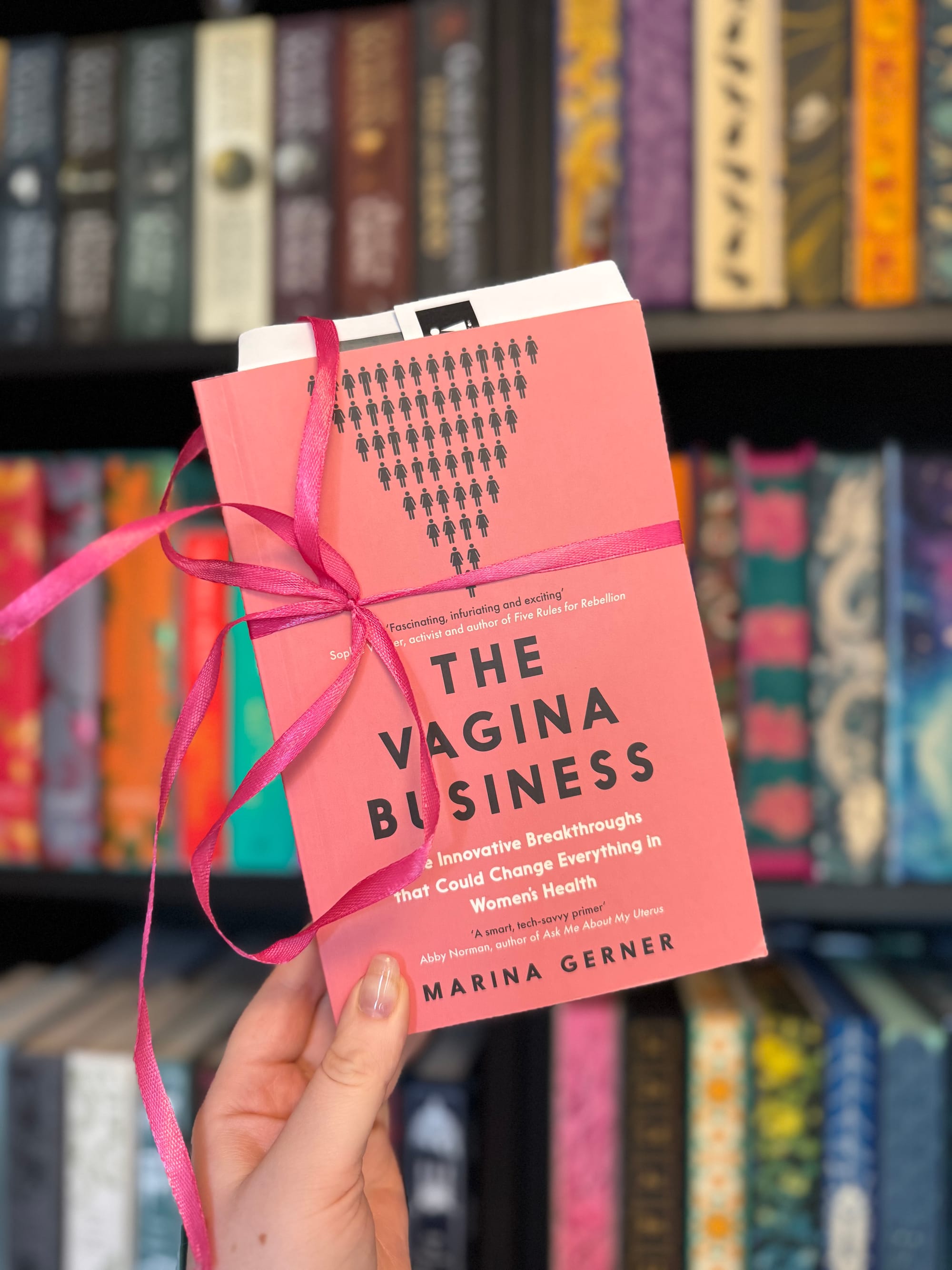 Hand holds a pink book titled "The Vagina Business," wrapped with a pink ribbon, against a colorful bookshelf background.