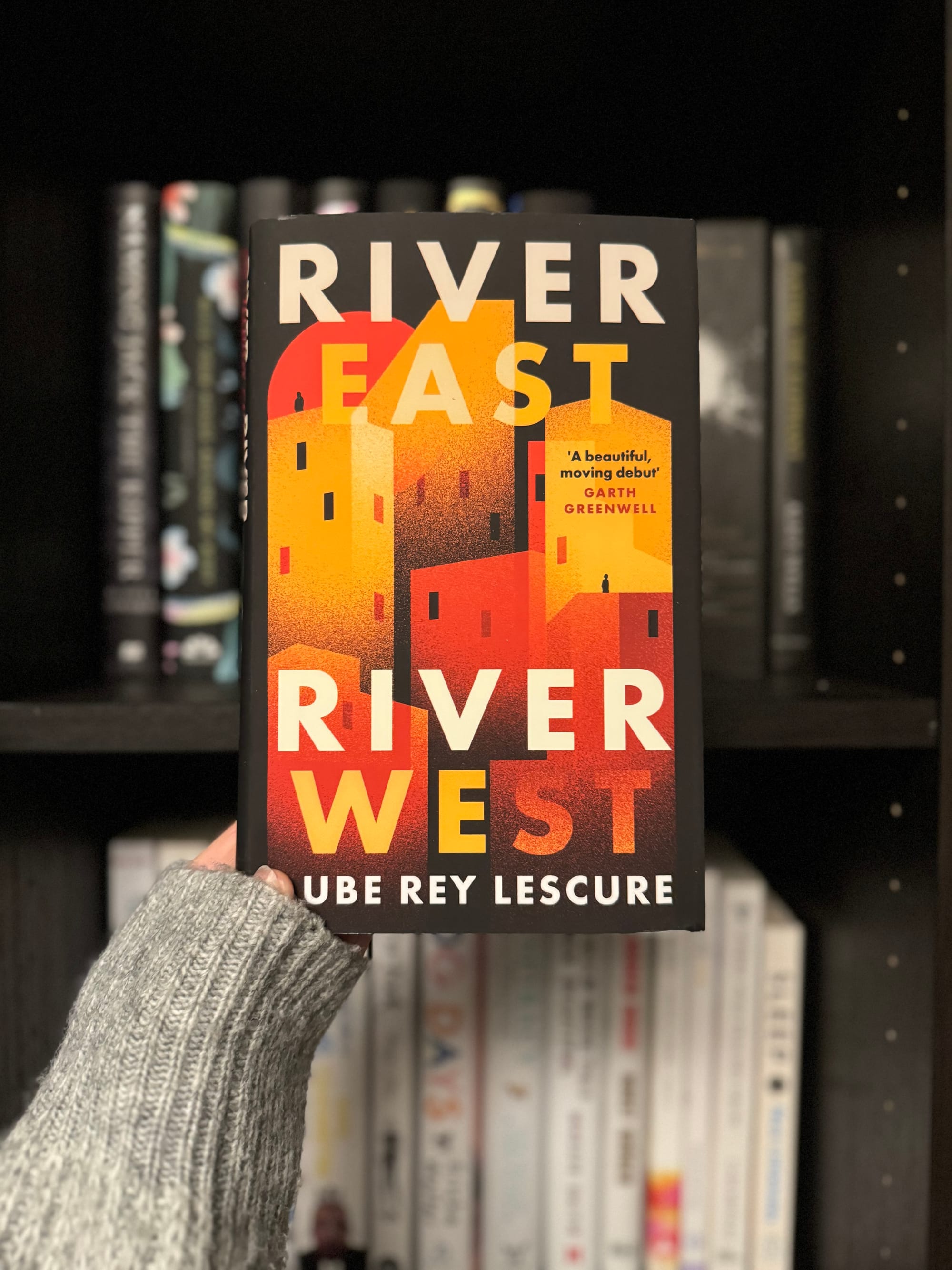 Hand holding a book titled "River East, River West" by Cube Rey Lescure, set against a book-filled shelf. Cover features red and yellow buildings.