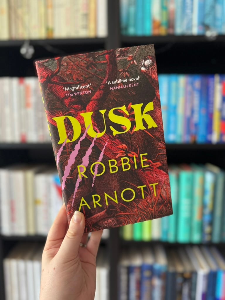 Hand holding "Dusk" book by Robbie Arnott with a vivid cover design. Background shows a blurred bookshelf with neatly arranged books.